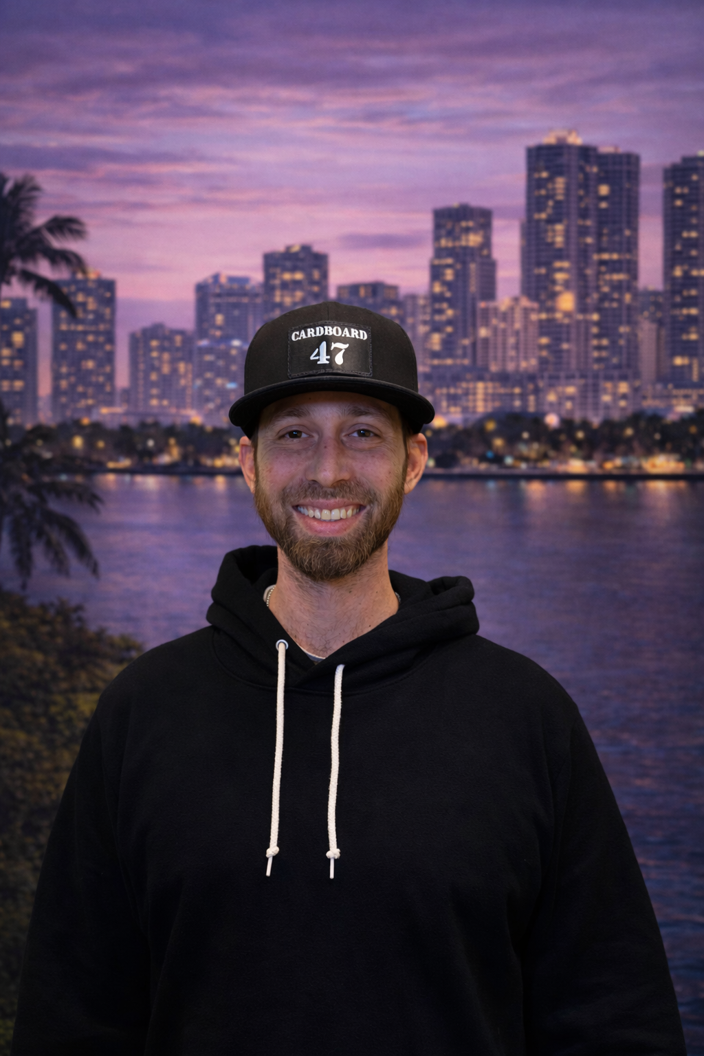 A smiling man with a beard and mustache wearing a black hoodie and a black cap with 'Cardboard 47' written on it. Behind him is a backdrop of a city skyline at dusk with high-rise buildings and water reflecting the purple and pink sky.