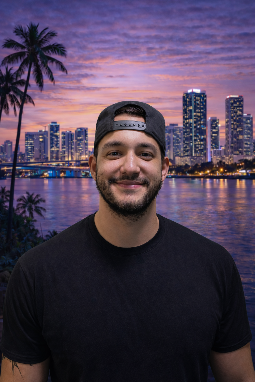 A young man with a beard and mustache wearing a black cap backwards and a black t-shirt, smiling in front of a city skyline at sunset with tall buildings, palm trees, and a body of water.