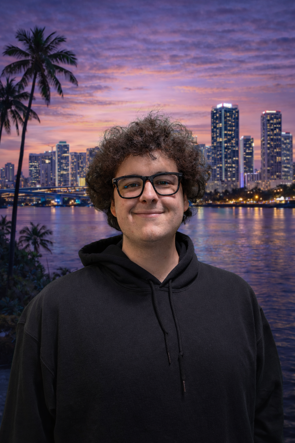 A young man with curly hair and glasses smiling in front of a city skyline at sunset, with tall buildings, palm trees, and a river reflecting lights.