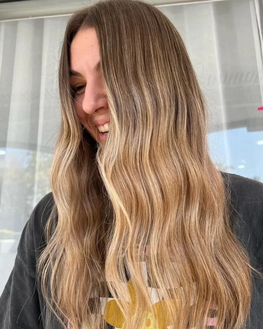 A woman with long, wavy, light brown hair smiling and looking down.