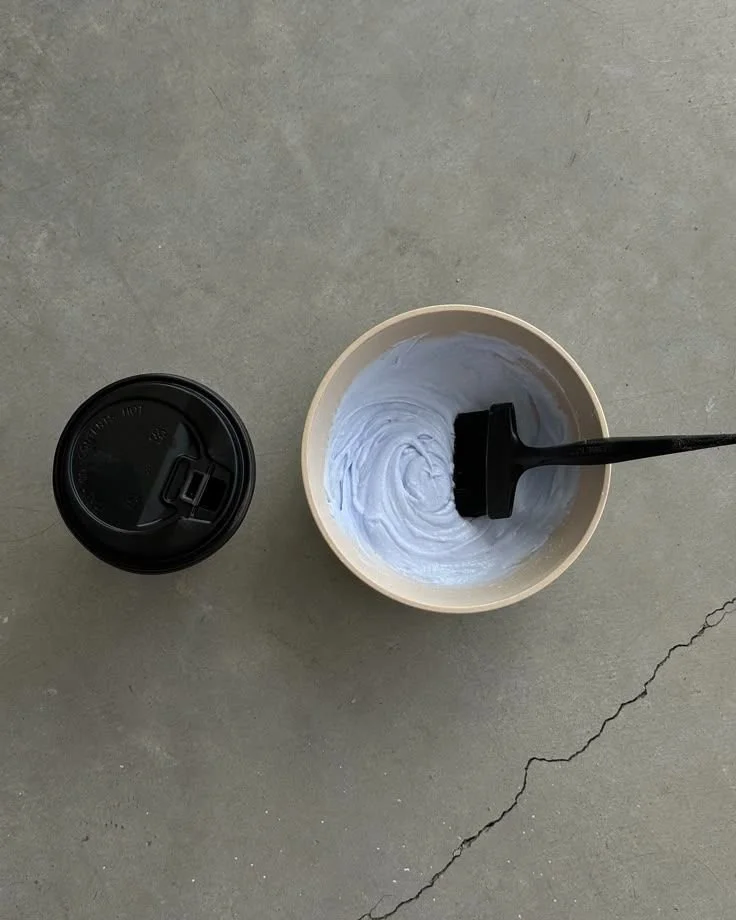 A white paper cup filled with a grayish substance and a black brush inside it, placed on a concrete surface. To the left is a black travel coffee mug.