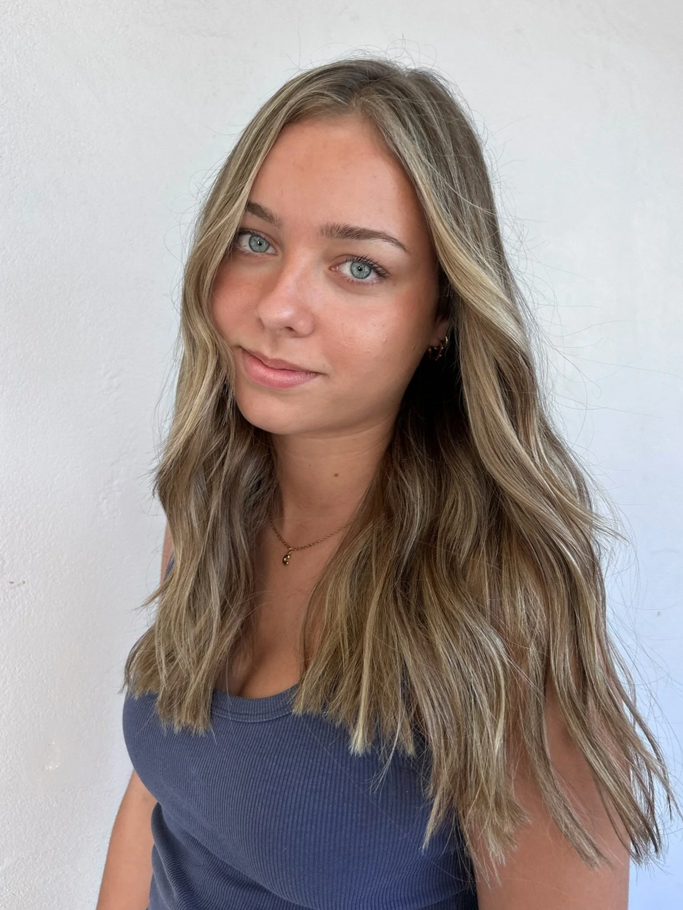 A young woman with long, wavy blonde hair and blue eyes, wearing a blue sleeveless top, a small necklace, and earrings, standing against a white background.