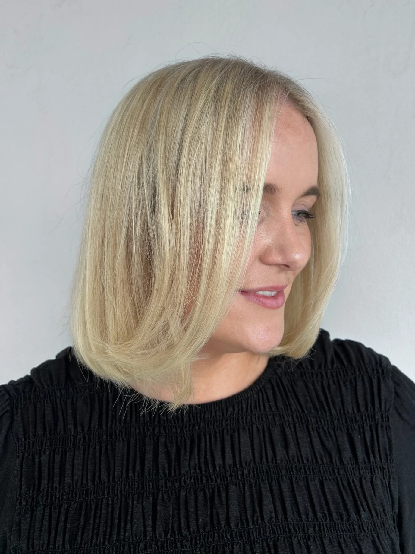 A woman with shoulder-length blonde hair, smiling softly, wearing a black textured top, standing against a plain light-colored background.