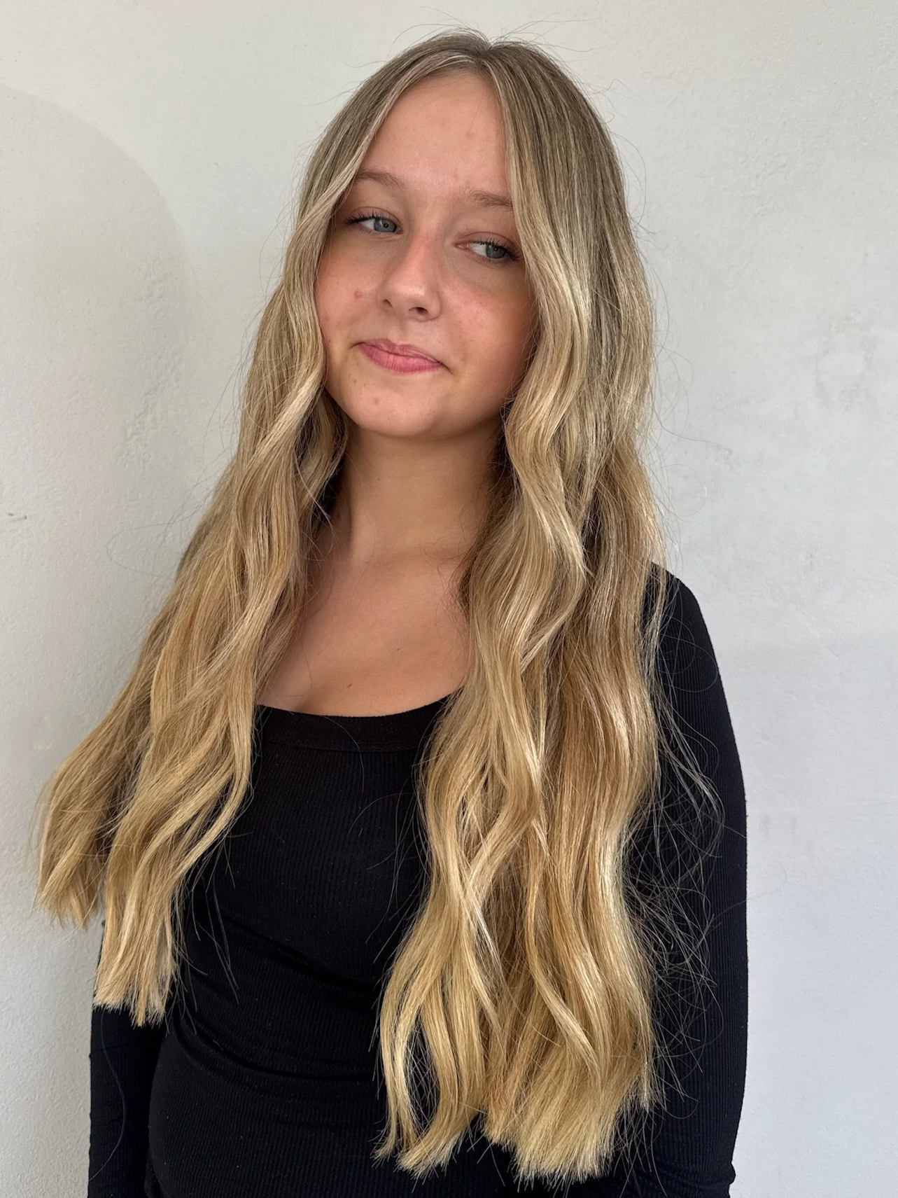 Young woman with long wavy blonde hair wearing a black top standing against a light-colored wall.