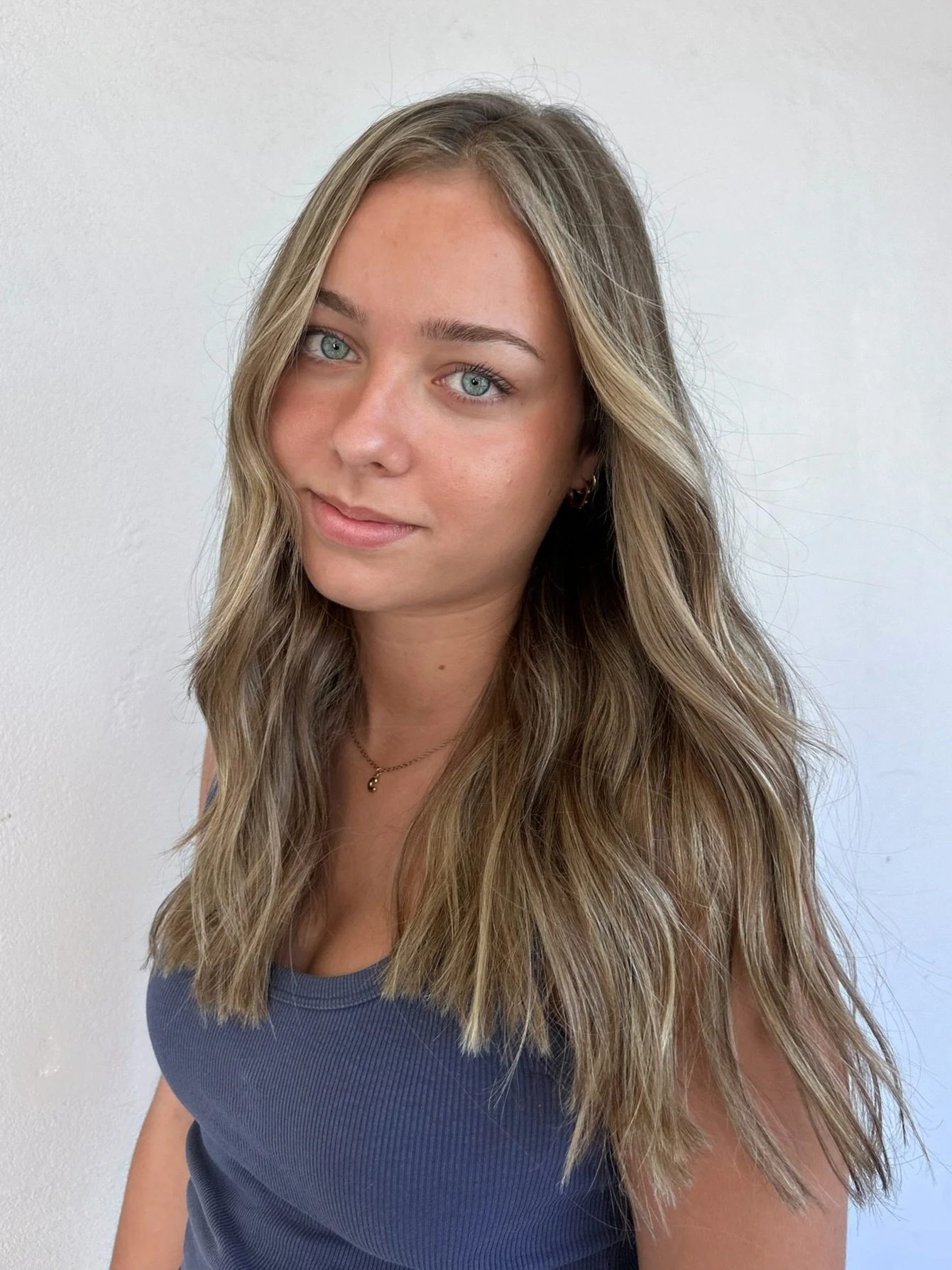 A young woman with long wavy blond hair and blue eyes, wearing a sleeveless blue top and a delicate gold necklace, standing against a plain white wall.