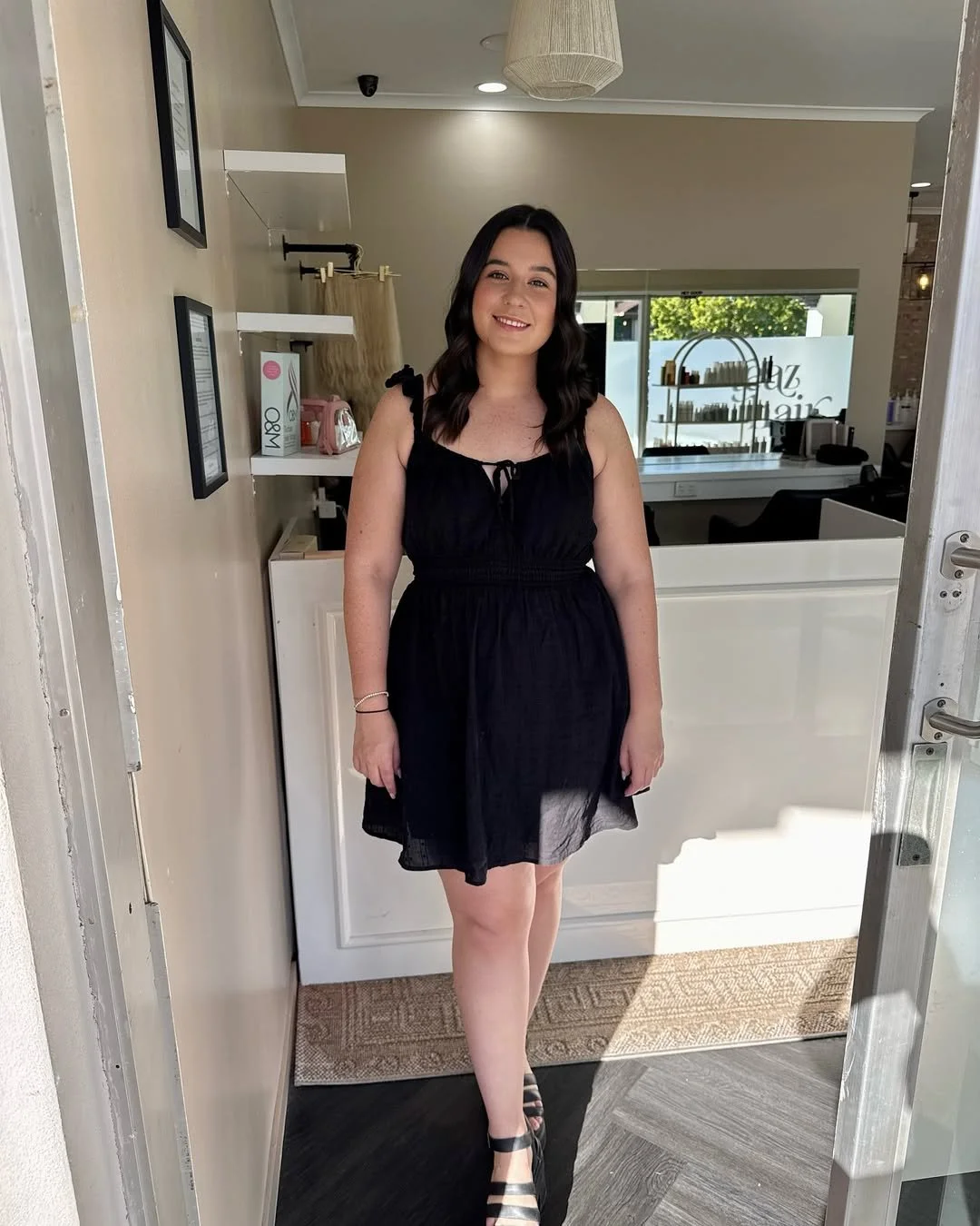 A woman with dark wavy hair wearing a black dress and striped heels standing inside a salon or spa, smiling at the camera.