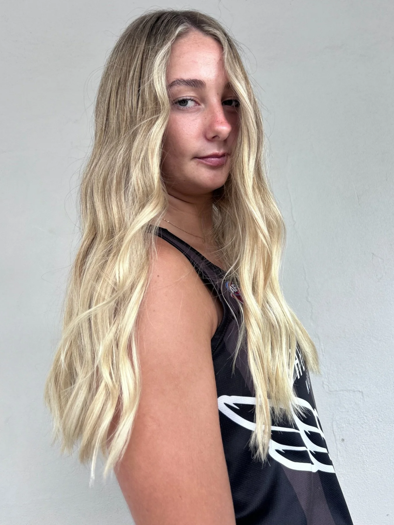 A young woman with long, wavy blonde hair, wearing a black sports jersey, standing against a plain, light-colored wall.