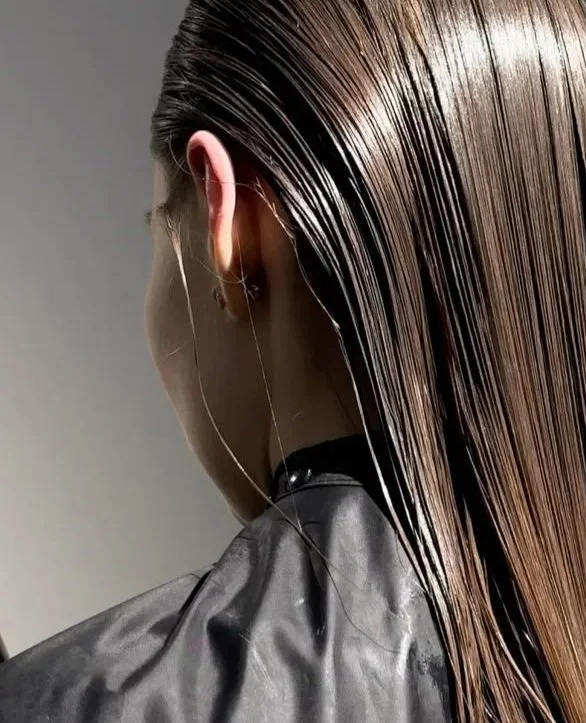 Close-up of a woman with wet, straight, brown hair, wearing a black jacket and earbuds. The photo captures the back and side of her head.