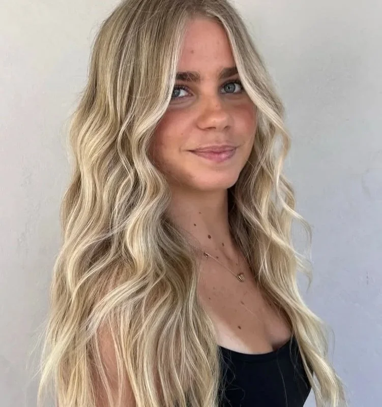 A woman with long, wavy blonde hair and blue eyes, wearing a black top and a delicate gold necklace, smiling against a plain light-colored background.
