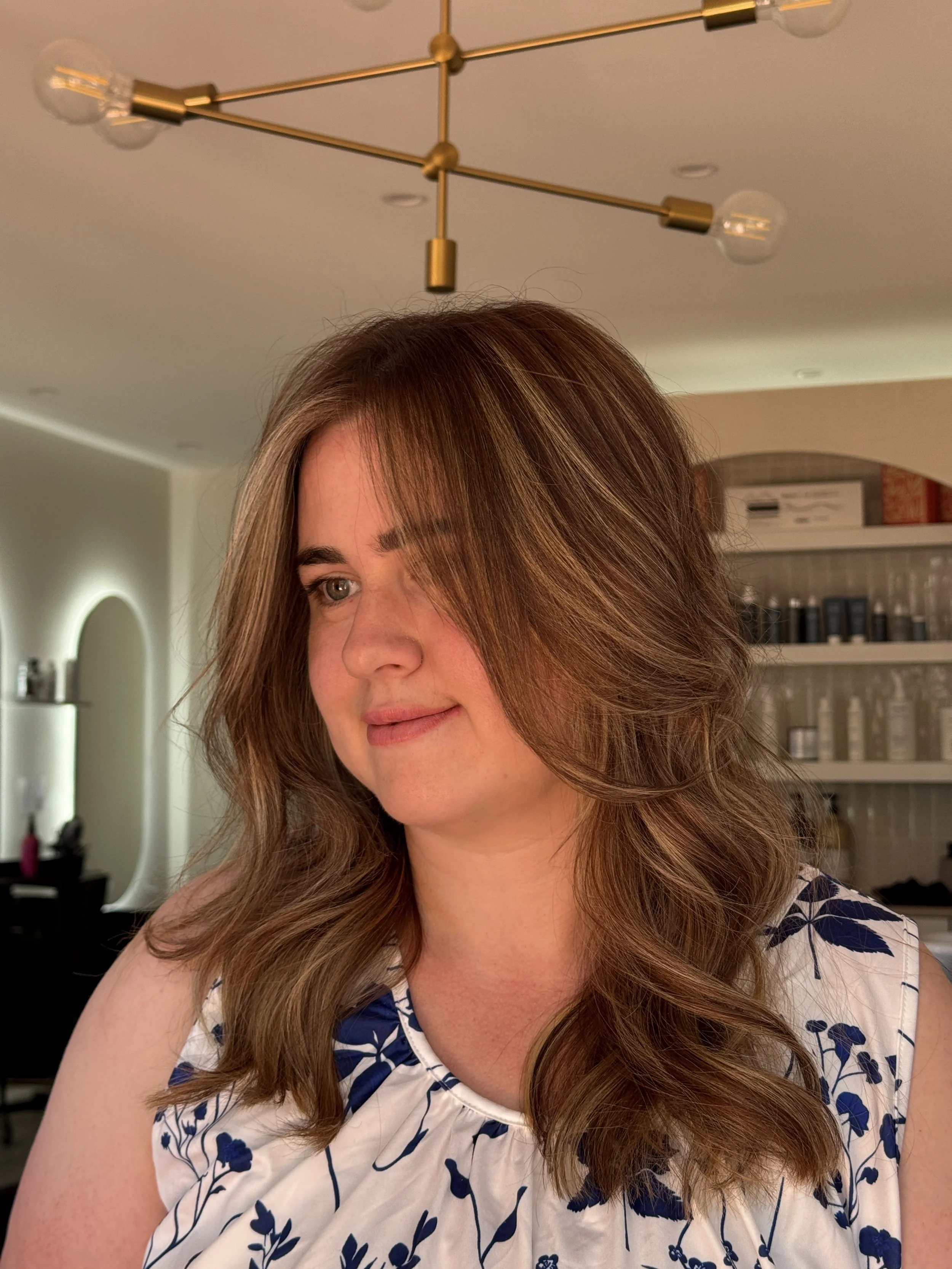 A woman with wavy, shoulder-length light brown hair, wearing a sleeveless white top with a dark blue floral pattern. She is in an indoor setting with warm lighting and a modern ceiling light fixture visible.