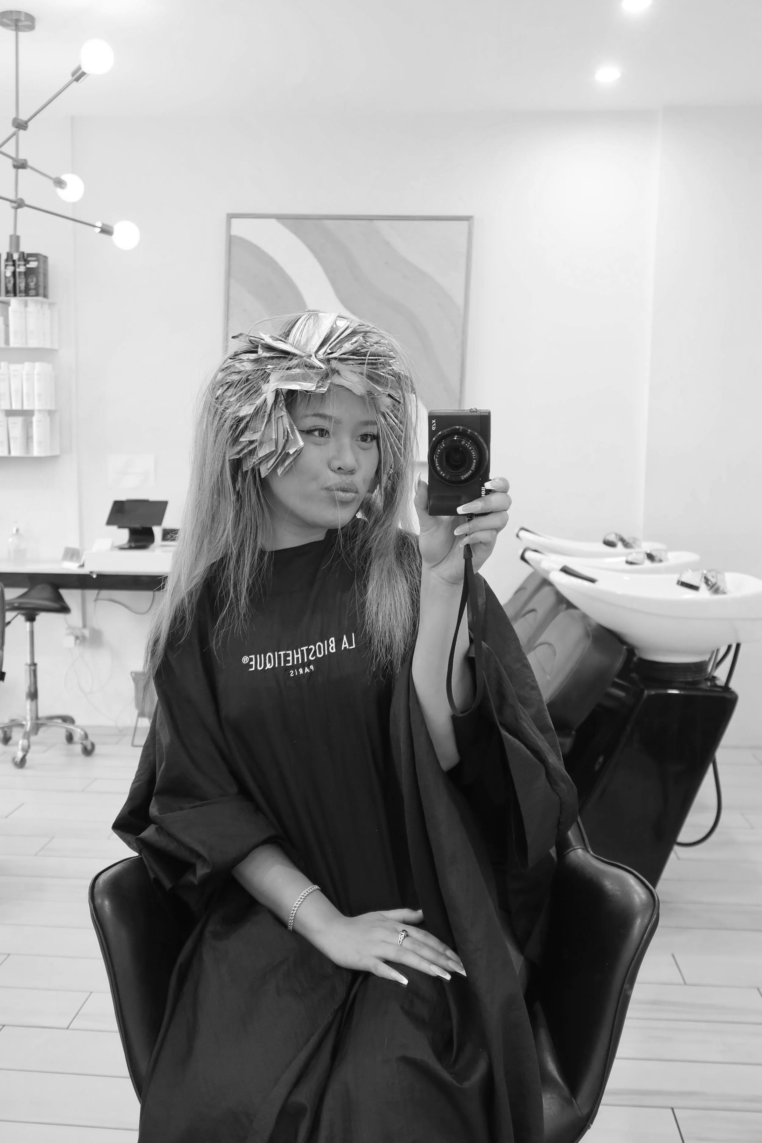A woman in a salon chair wearing a haircutting cape, with hair rollers in her hair, taking a selfie in a salon mirror. The salon has hair washing sinks and shelves with products in the background.