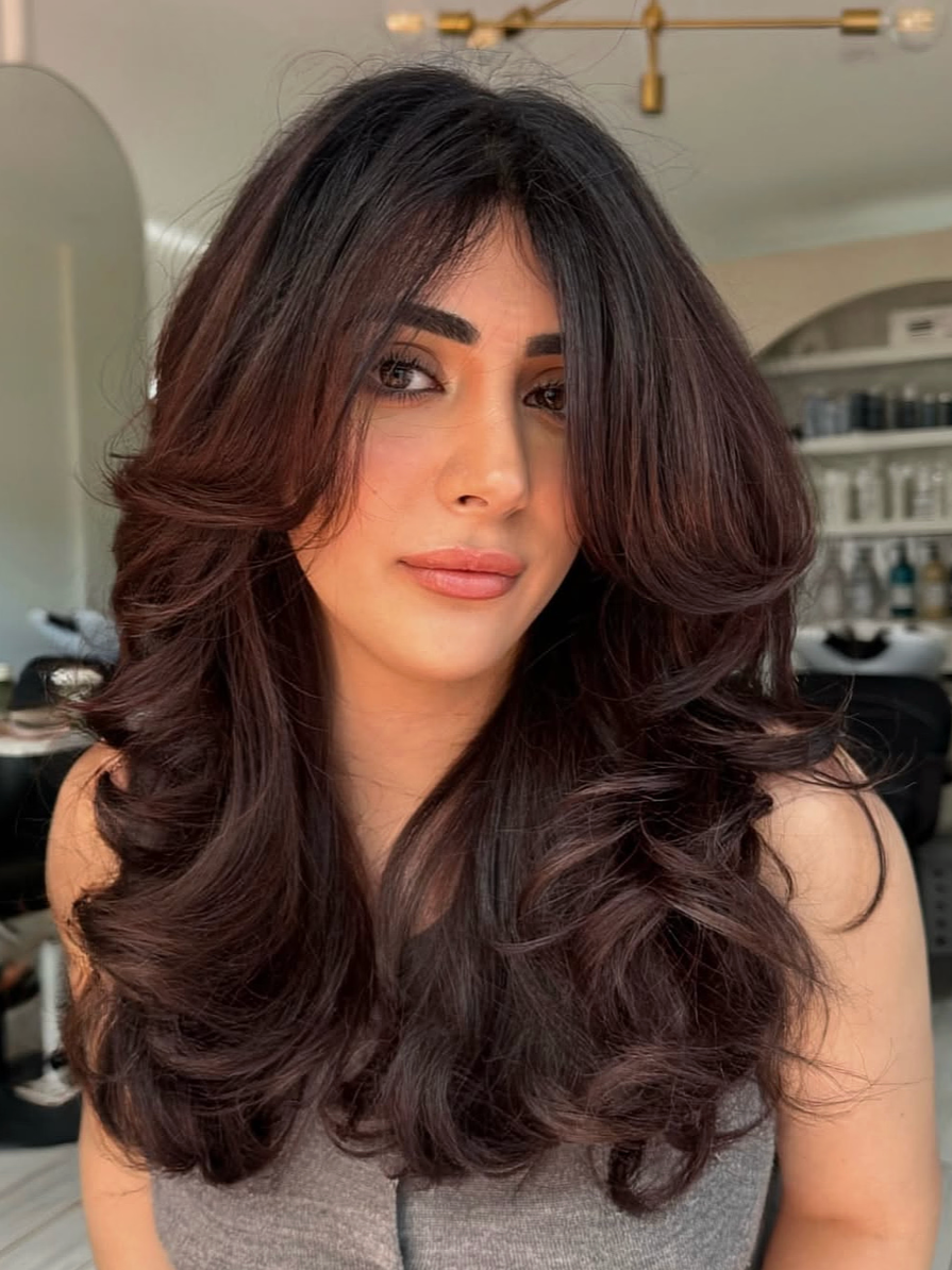 Woman with long, voluminous dark brown hair styled in loose curls, wearing a sleeveless top, inside a salon with shelves of hair products in the background.