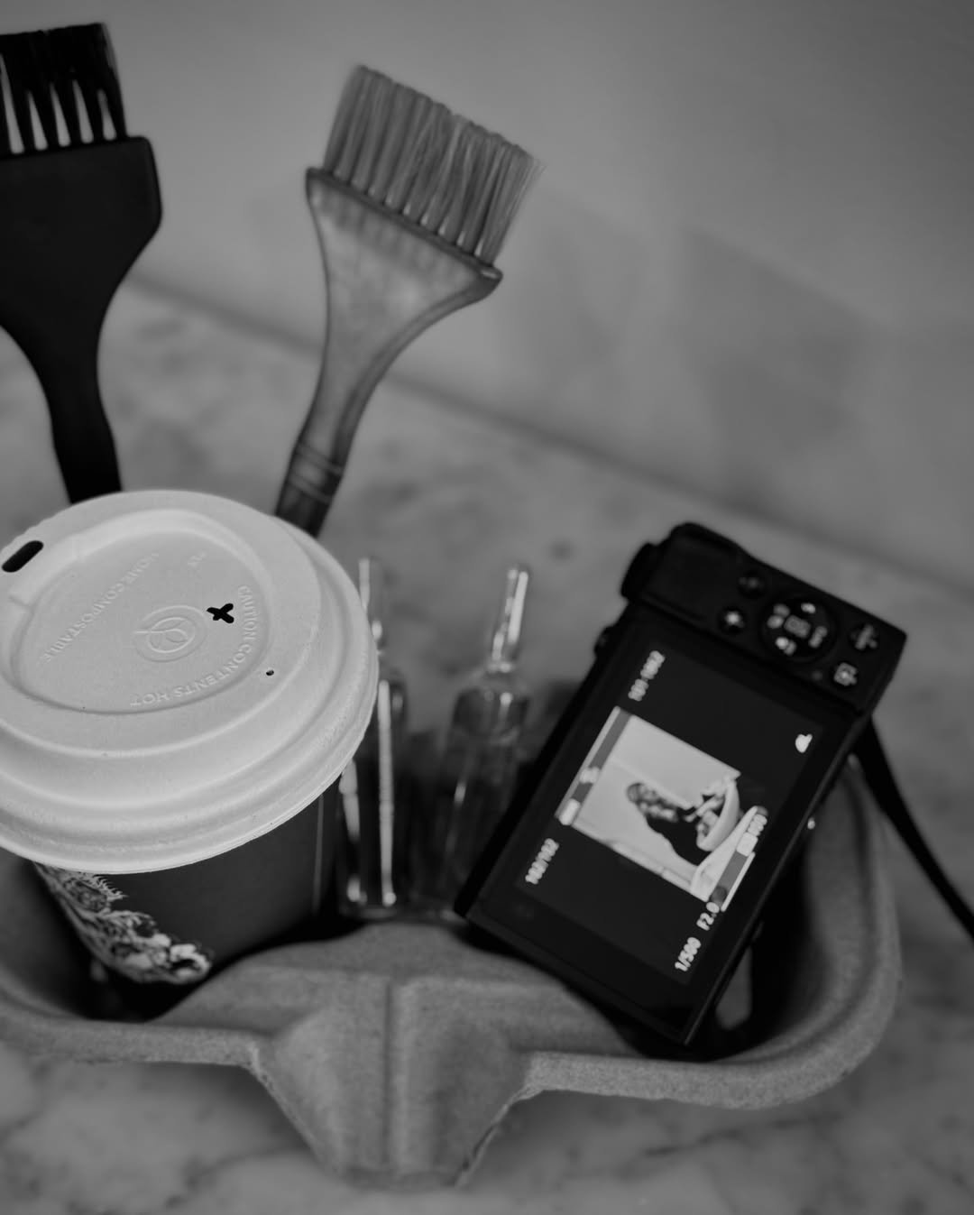 Black and white photograph of a table with a coffee cup, two paintbrushes, a small glass bottle, and a camera showing a woman posing for a photo.