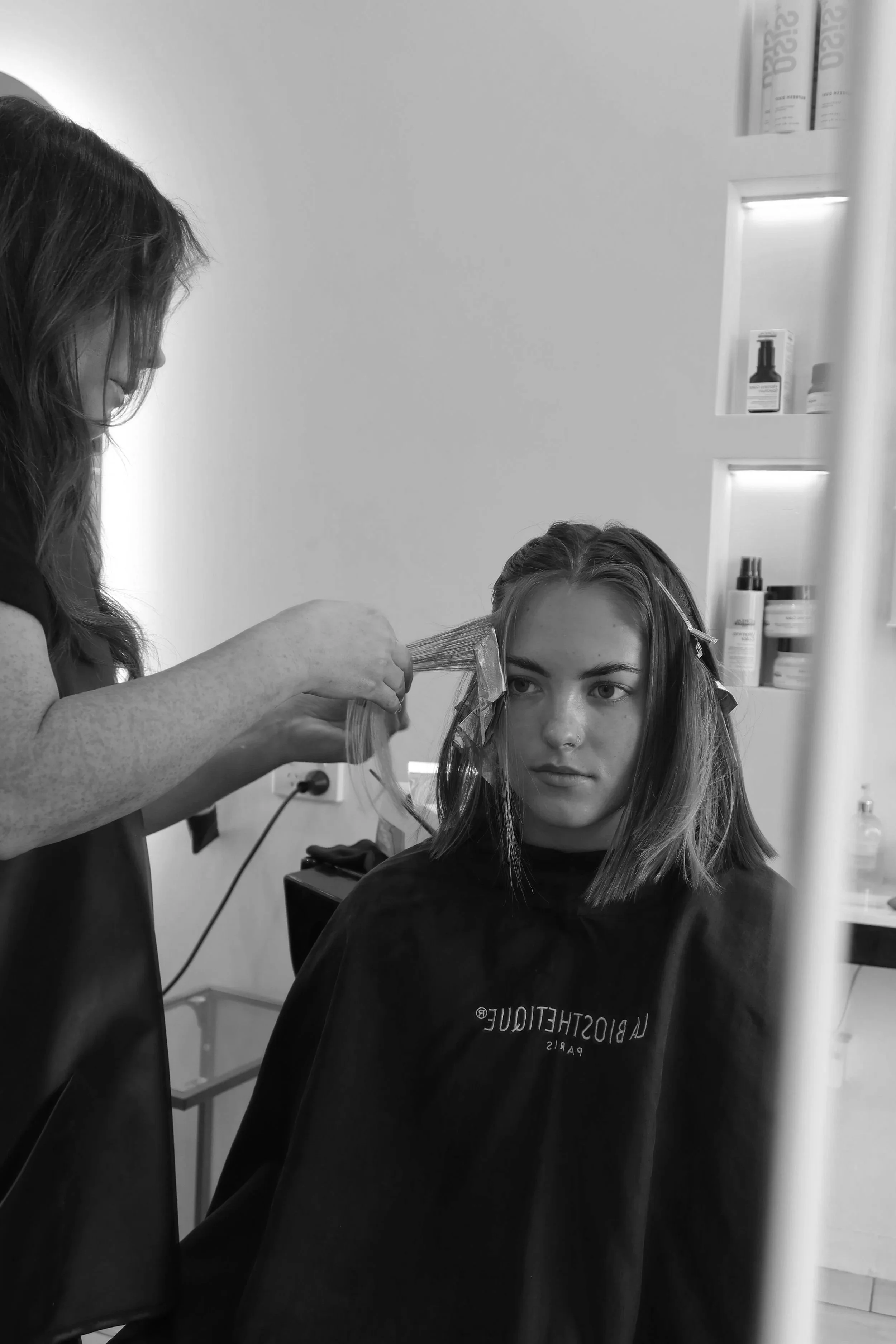 A woman at a salon getting her hair styled by a hairstylist. The stylist is working on her hair with rollers in a salon setting, with shelves of hair products in the background.