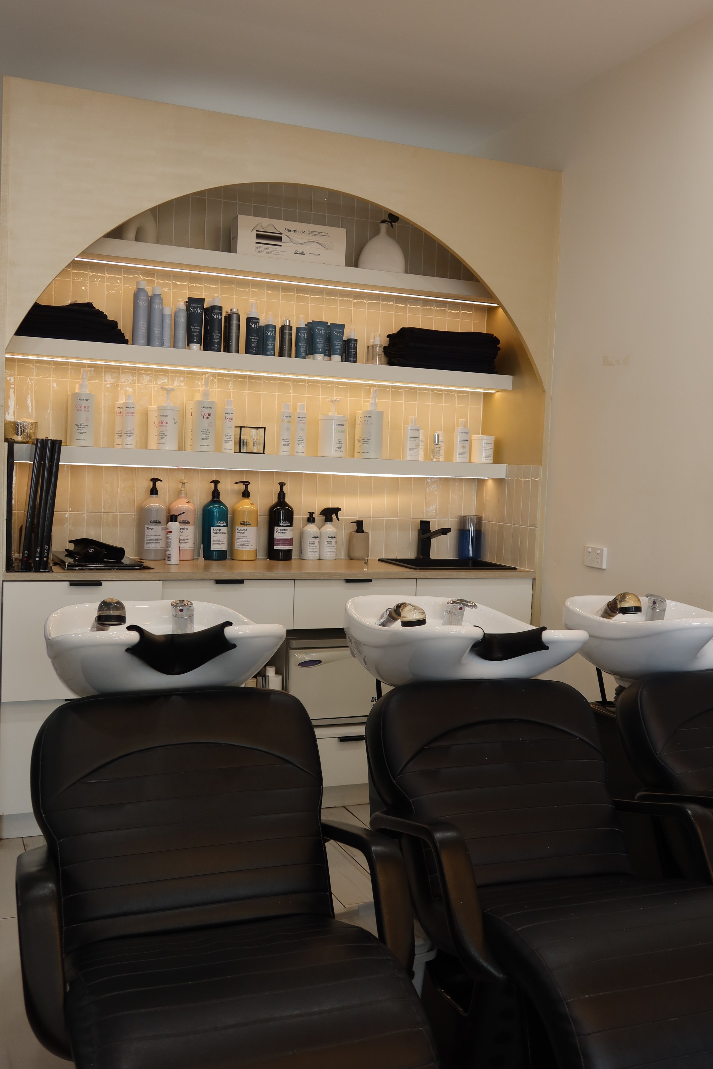 Inside a hair salon with black chairs, white shampoo bowls, and shelves displaying hair care products.