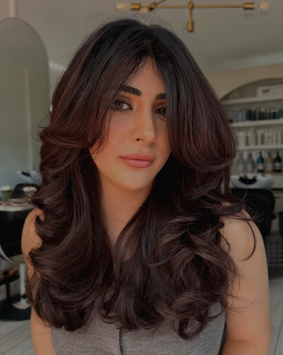 A woman with long, voluminous dark brown hair styled in loose curls, wearing a gray top, indoors with blurred background including shelves and a mirror.