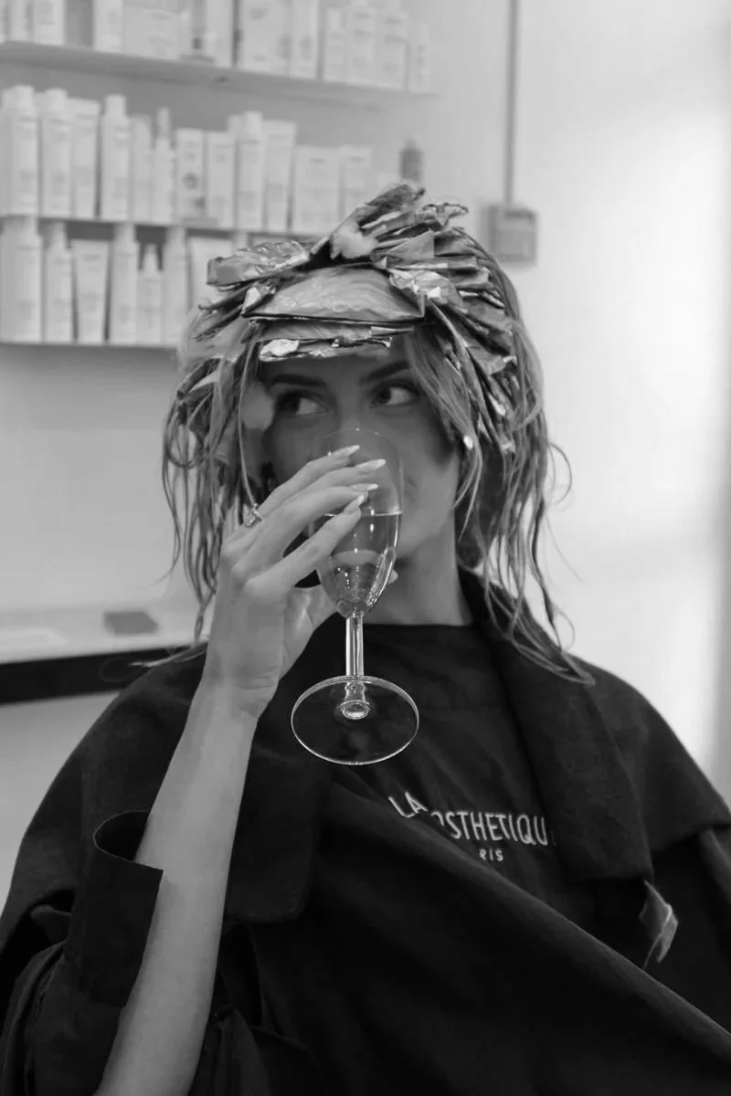 Person with hair wrapped in aluminum foil, holding a glass of champagne, sitting in a salon or hair treatment setting.