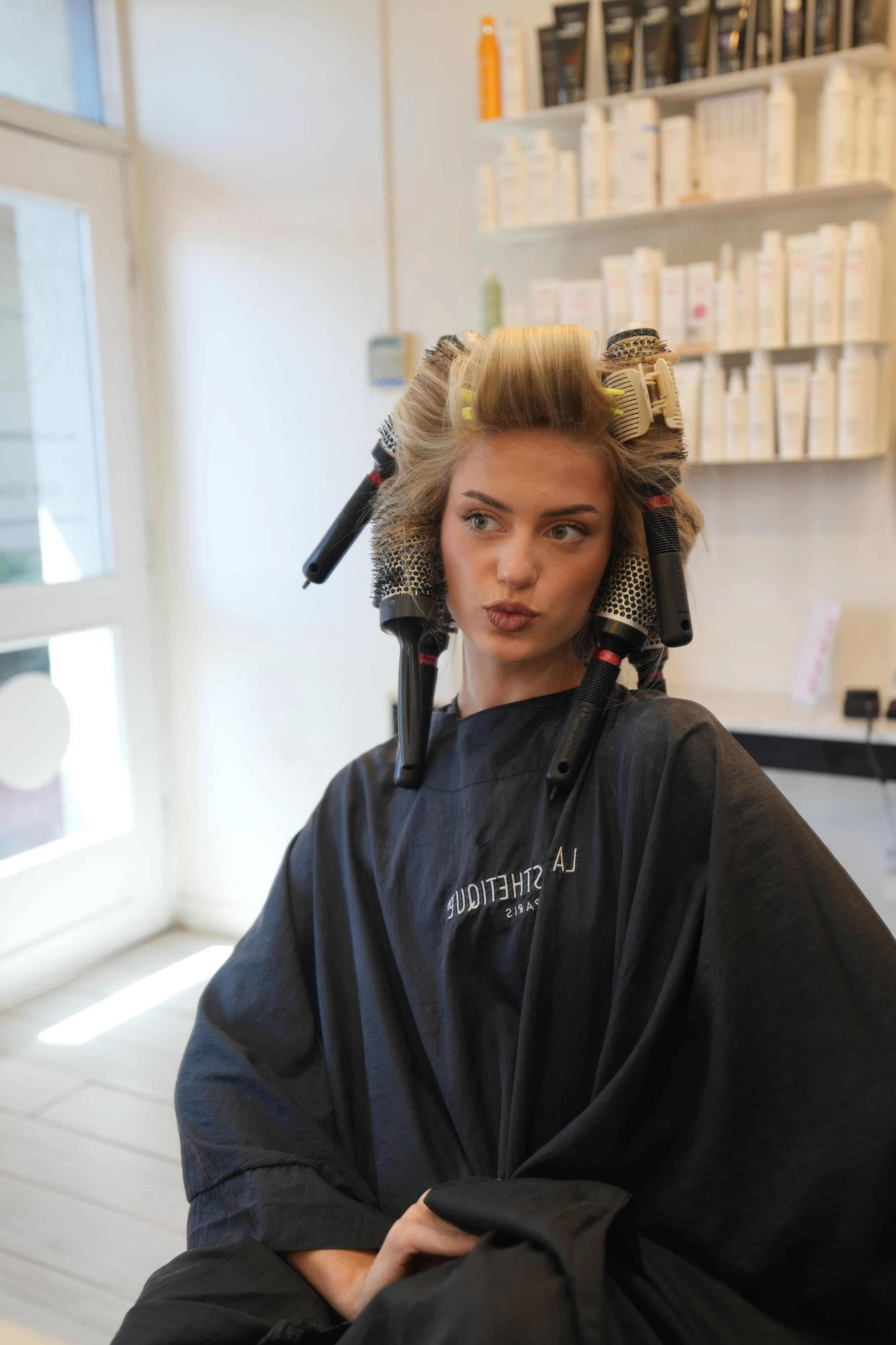 A woman with hairstyling rollers and curlers in her hair sitting in a salon chair, wearing a black salon cape, with a shelf of hair products behind her.