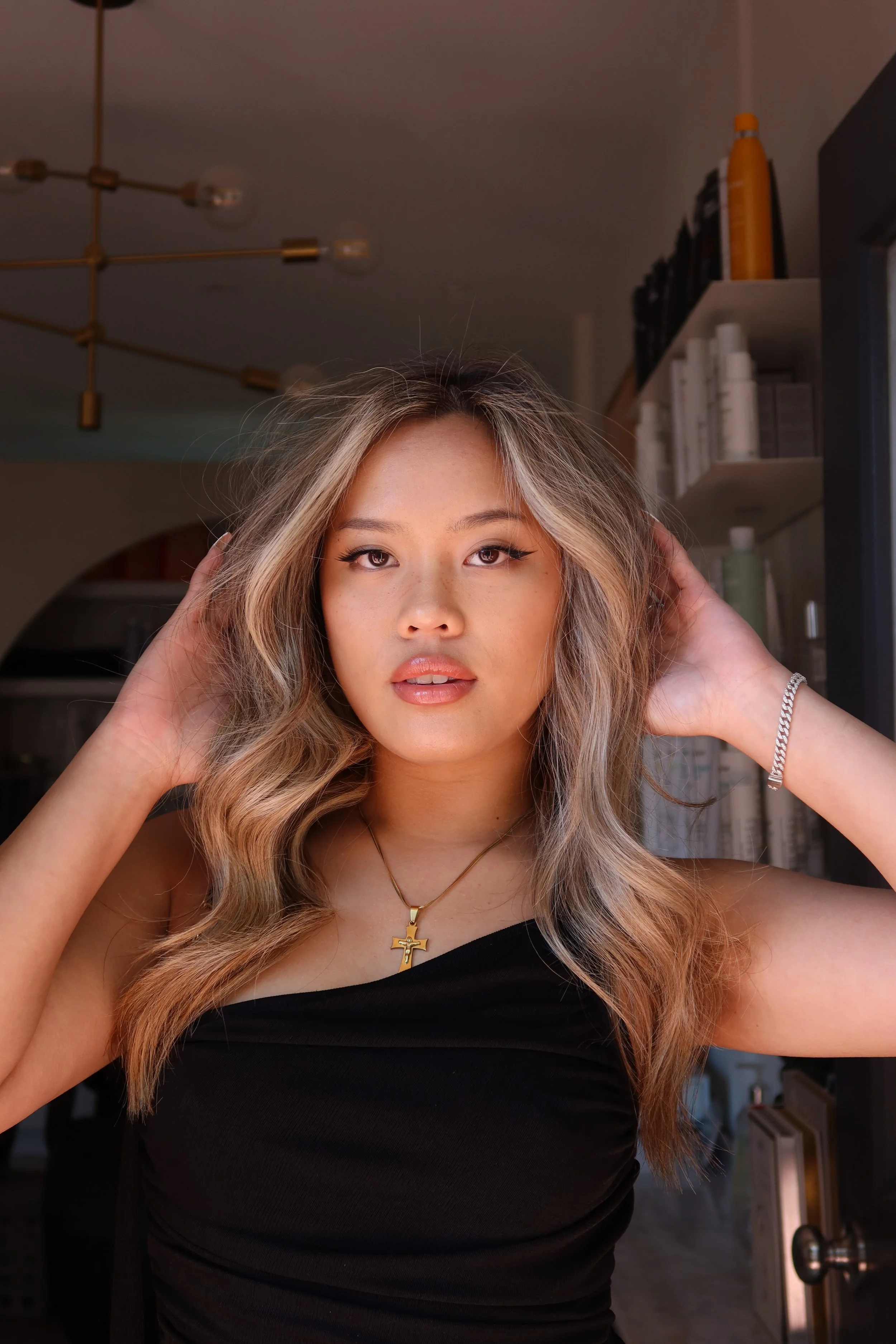 A young woman with wavy blonde hair, wearing a black strapless top, gold necklace with a cross, and silver bracelet, standing indoors.