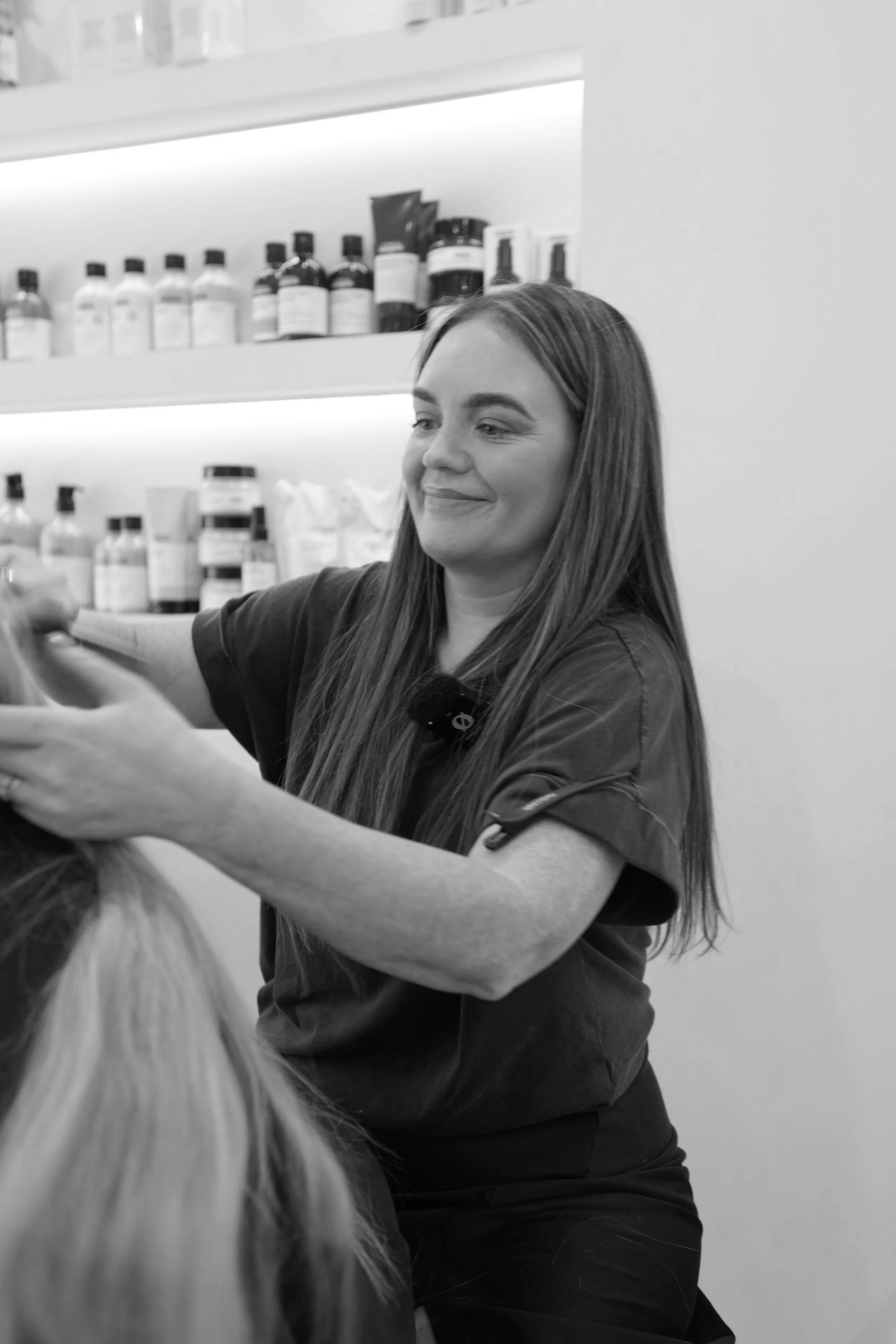 A woman with long hair styling a person's hair in a salon, with shelves of bottles and hair products in the background.