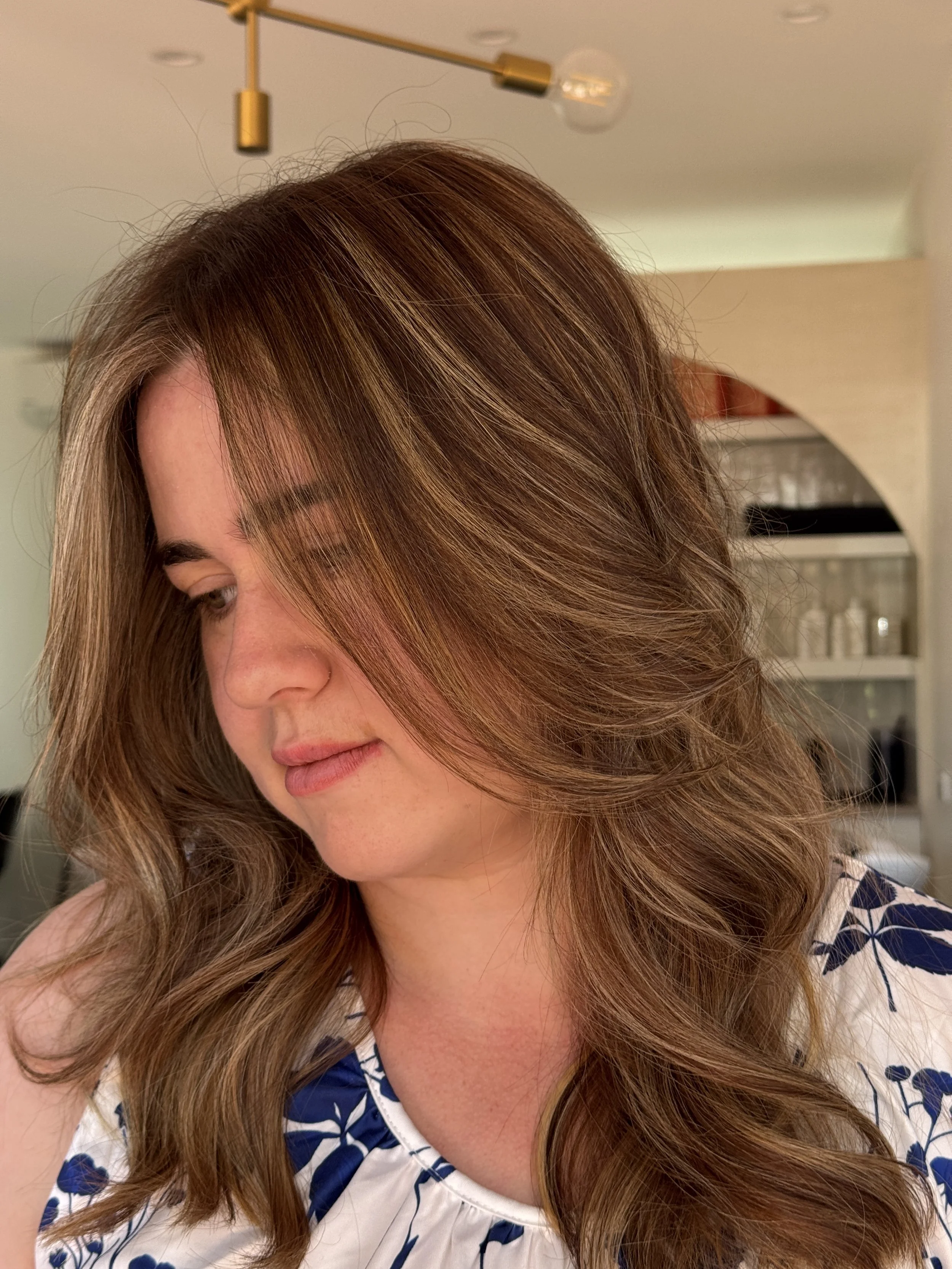 A woman with long, wavy brown hair with blonde highlights, wearing a white blouse with blue floral patterns, looking down.
