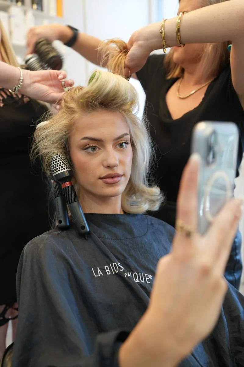 Woman getting her hair styled at a salon, taking a selfie with a smartphone.