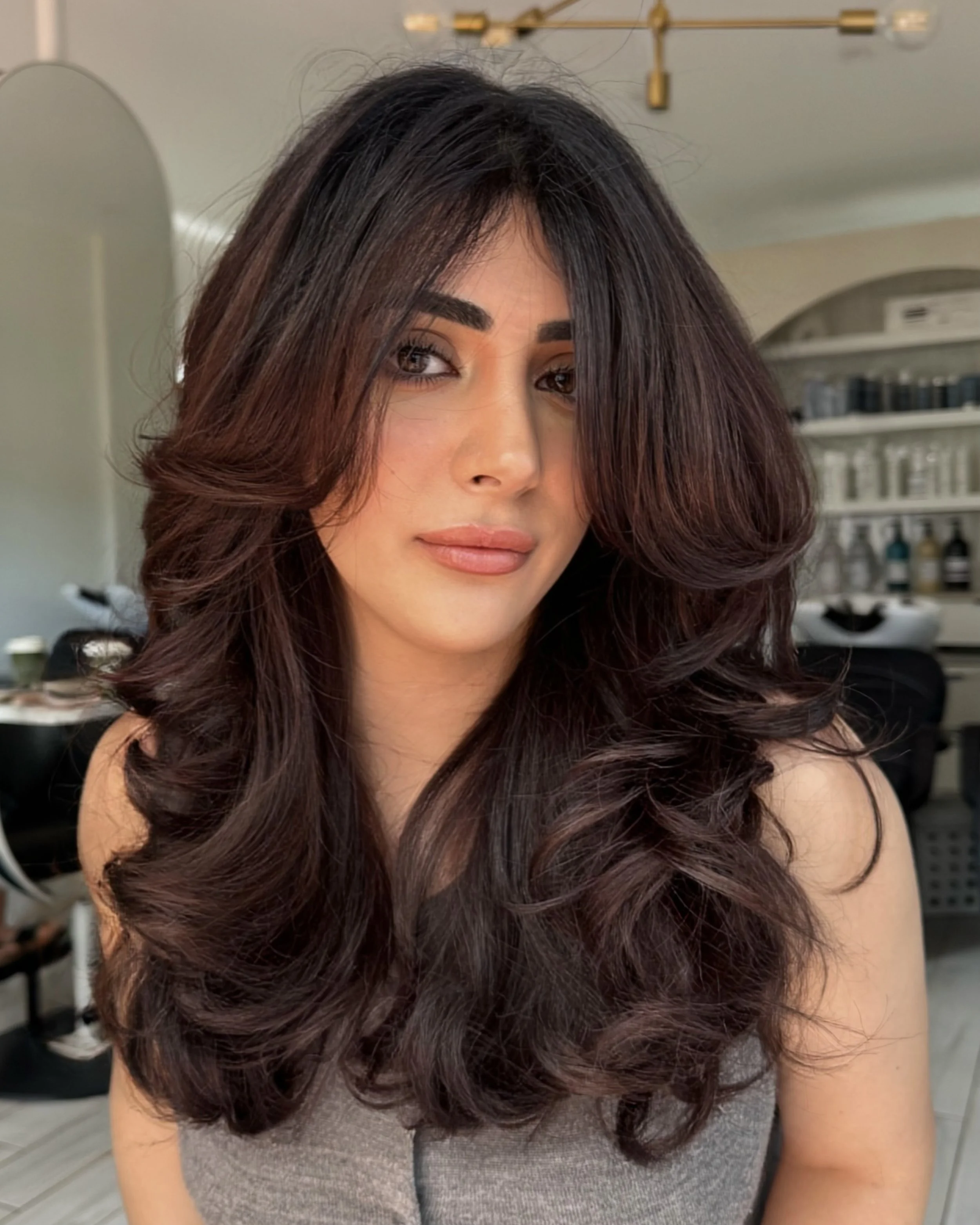 Woman with long, voluminous, wavy dark brown hair taking a selfie in a salon or home setting.