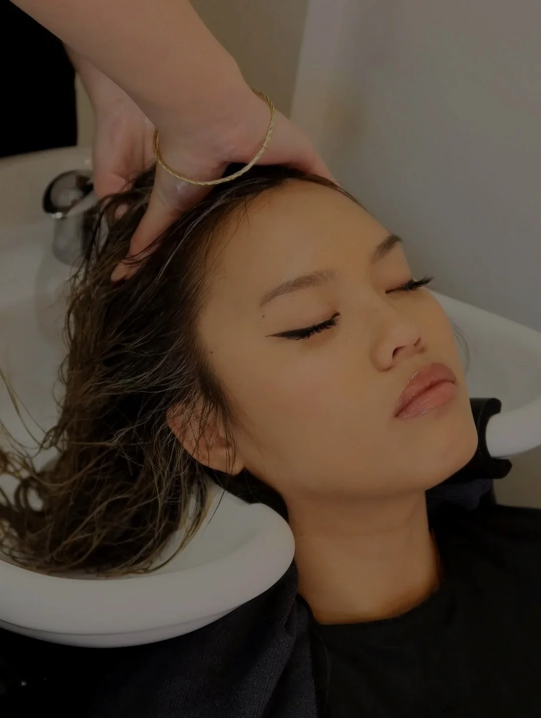 A woman lying back with eyes closed, receiving a scalp massage or treatment in a spa or salon setting.