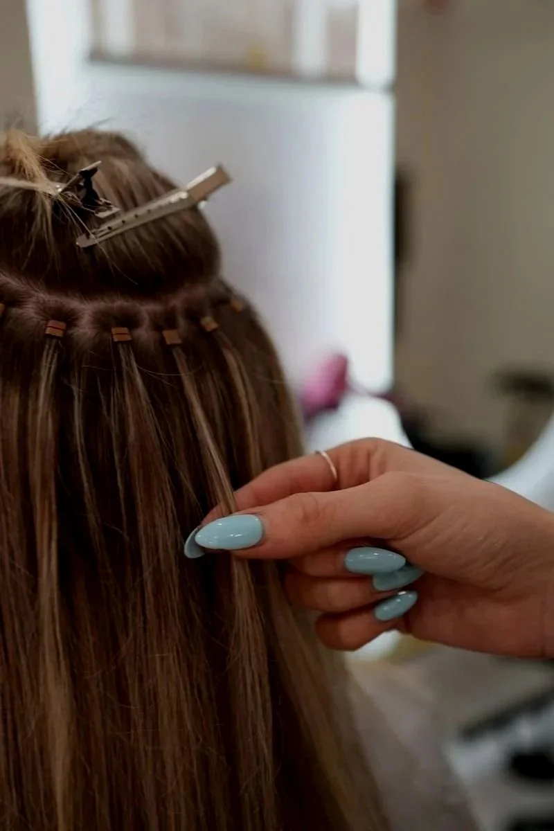 A hairstylist applying hair extensions to a person's brown hair with clip-in extensions.