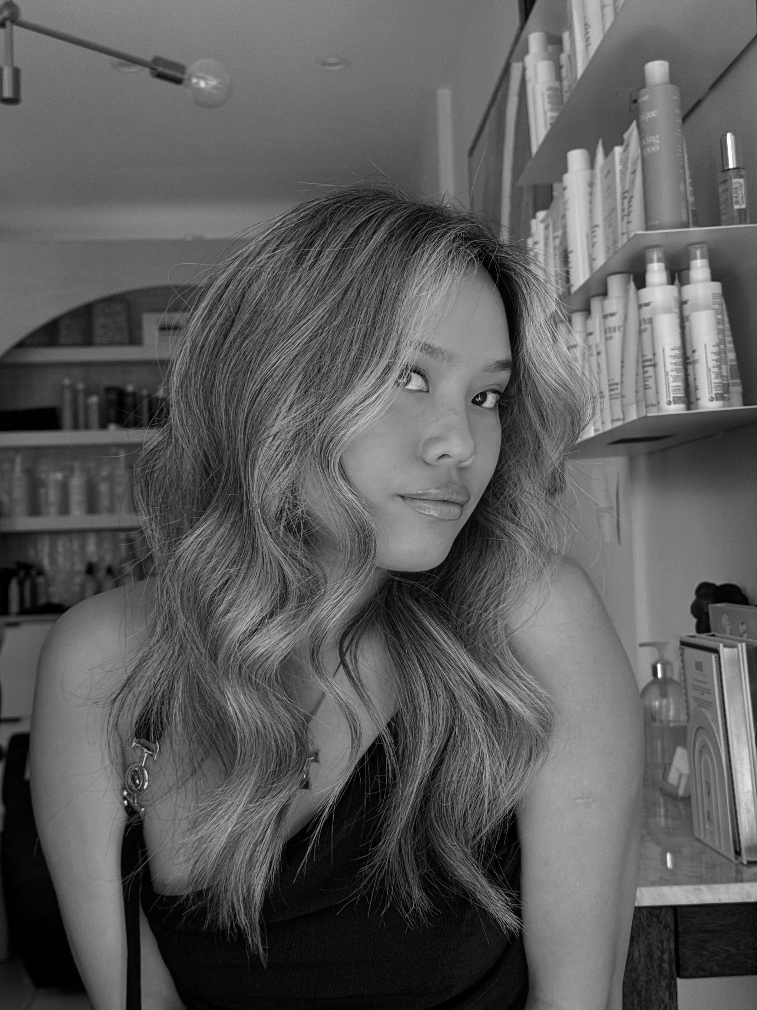 A woman with wavy hair sitting in a room with shelves of bottles and books around her.