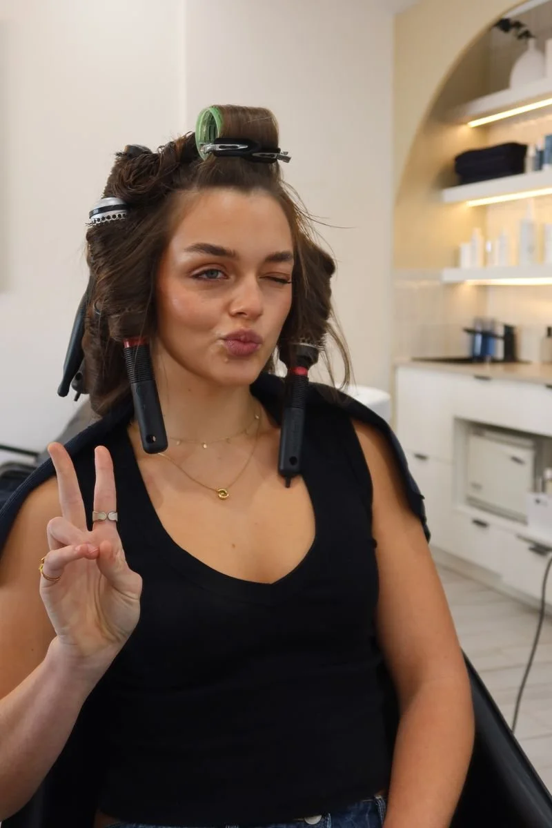 A woman with hair rollers and curlers in her hair, sitting in a hair salon, making a peace sign with her right hand and winking.