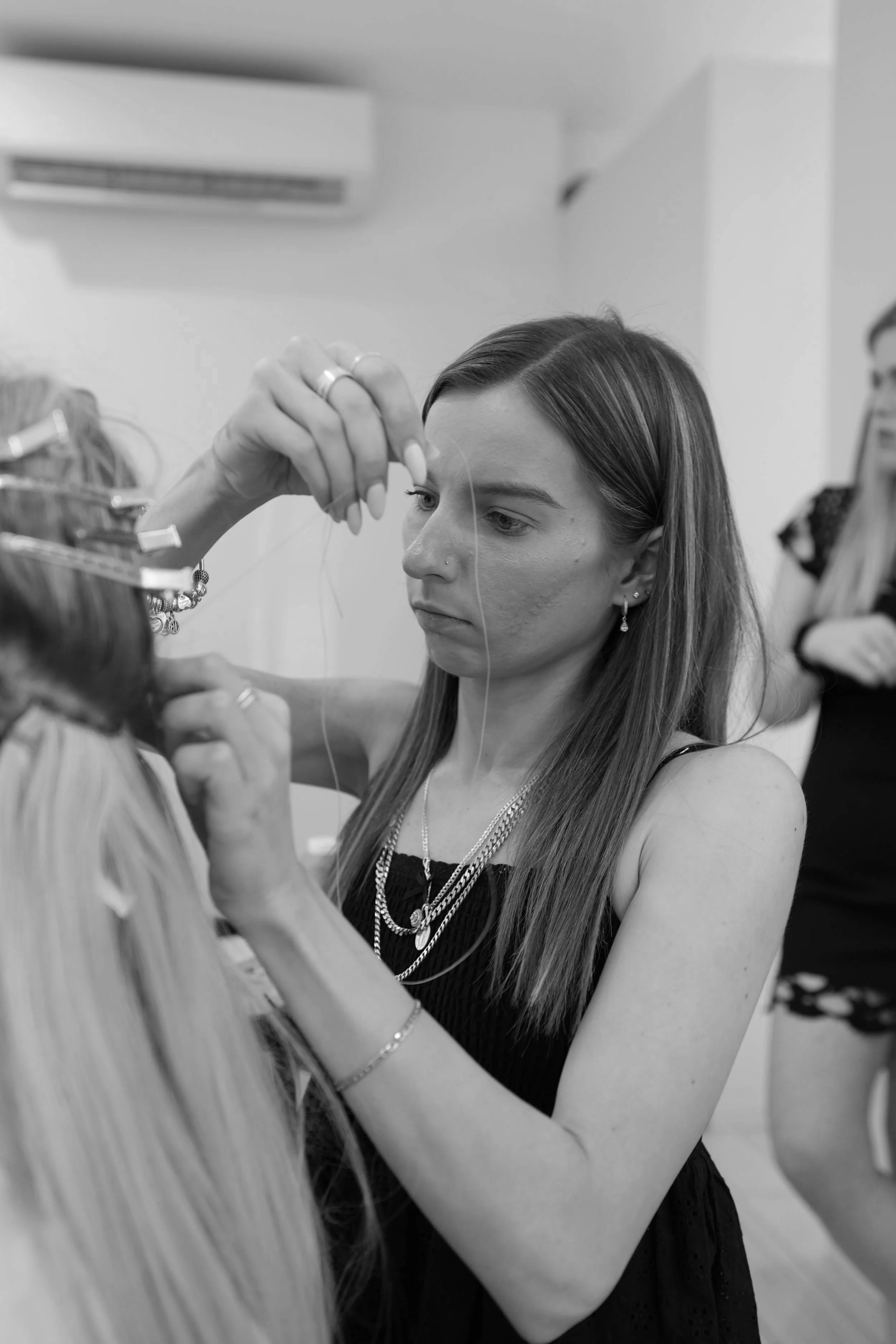 A woman with long hair and jewelry working on a client’s hair in a salon, with another person in the background.