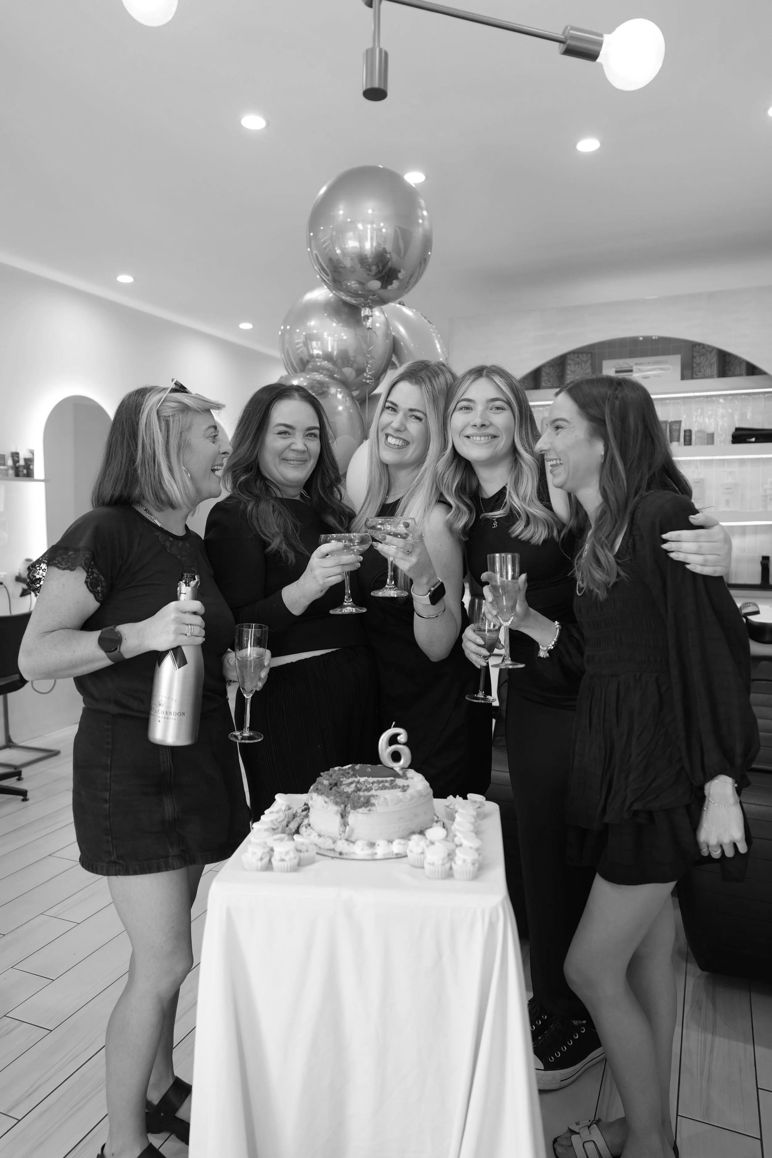 Black and white photo of five women celebrating birthday with champagne glasses and cake, with balloons in the background