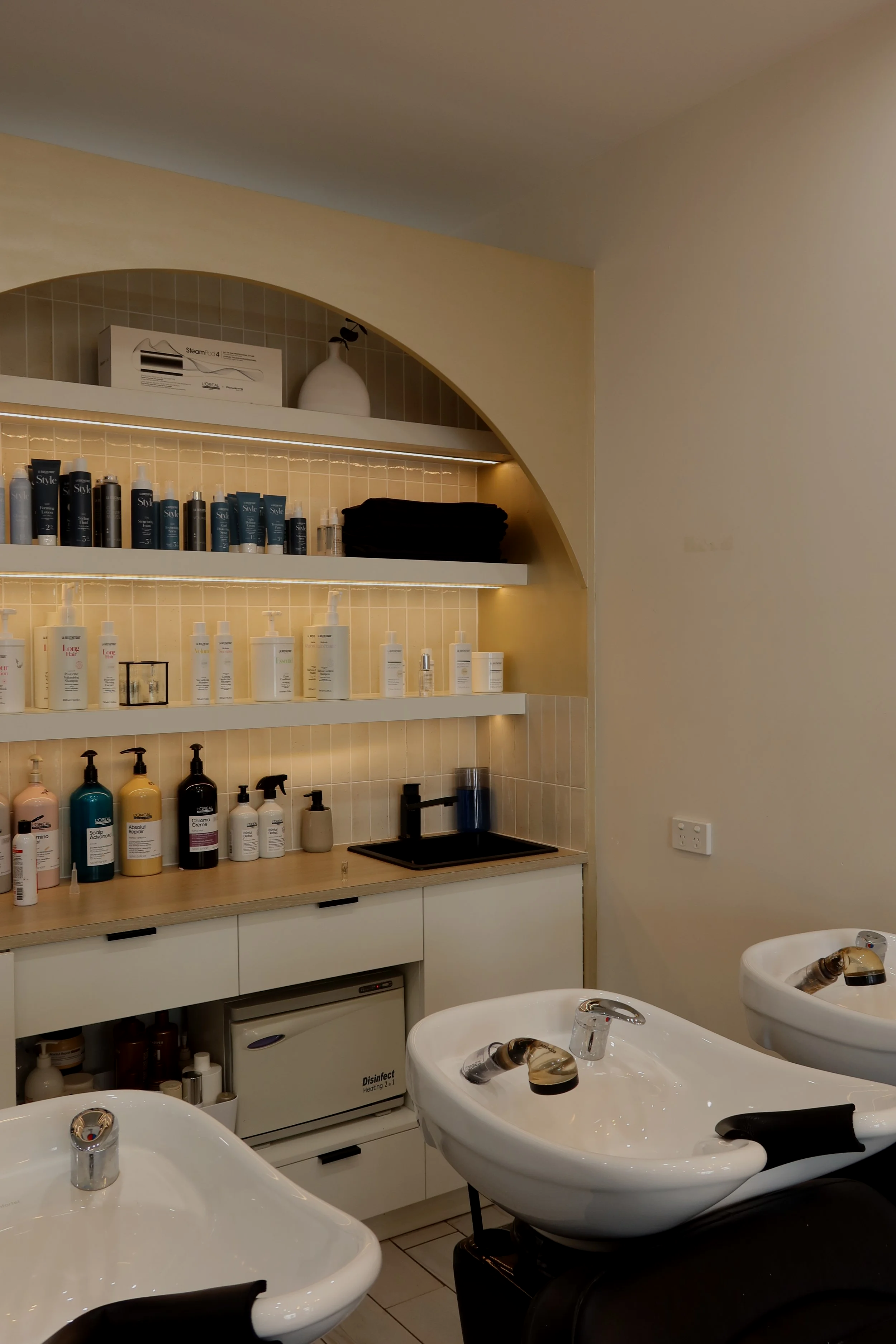 Hair salon or spa shampoo and treatment bowls with shelves of hair and skincare products, a small sink, and a countertop.