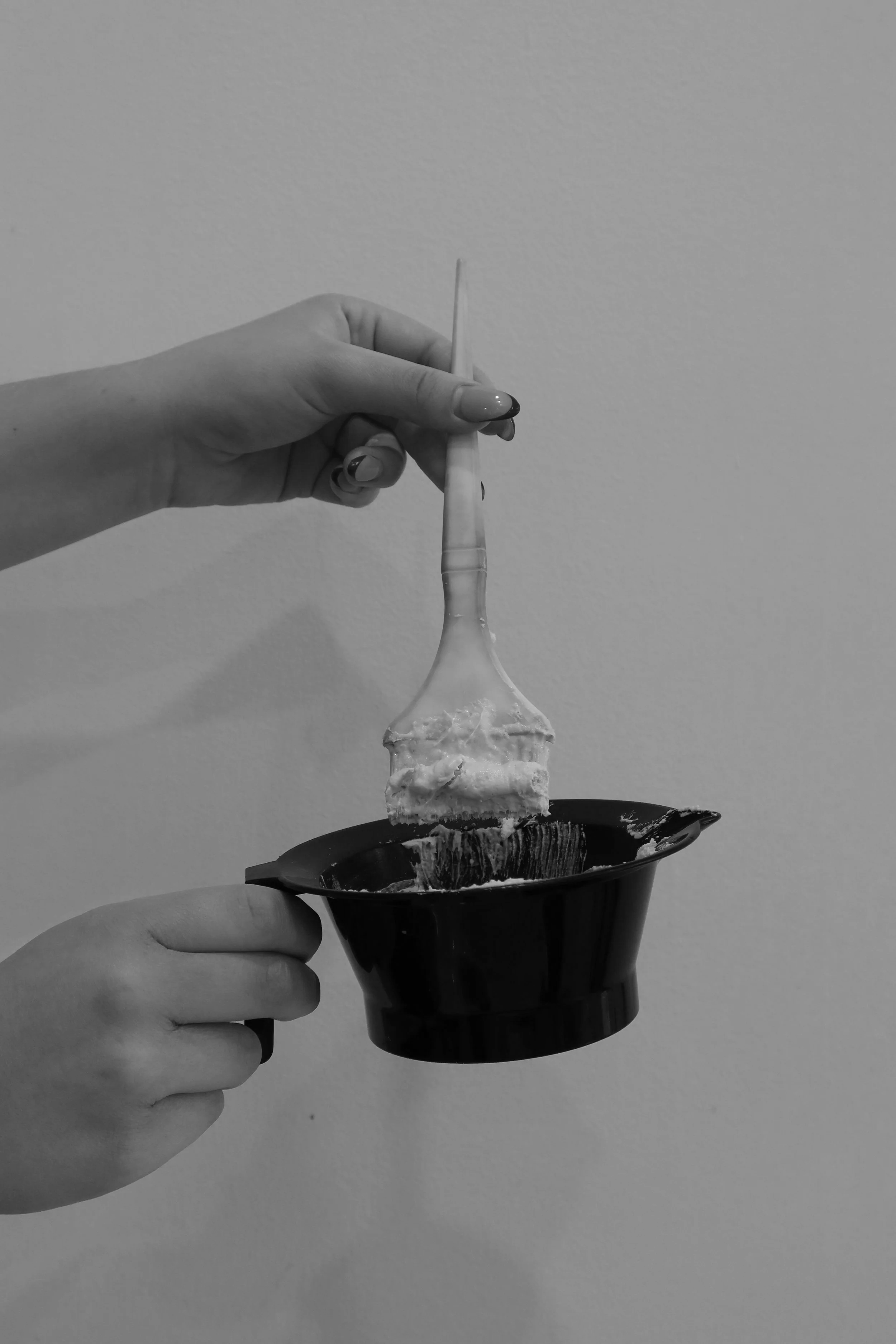 A person holding a toothbrush with a mixture of toothpaste and cream, above a black disposable bowl against a plain wall.