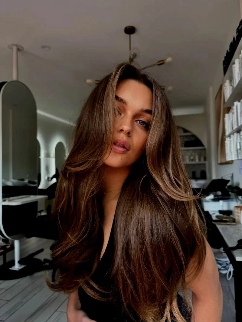 A woman with long, wavy brown hair and light skin looking at the camera indoors, with a modern interior in the background.
