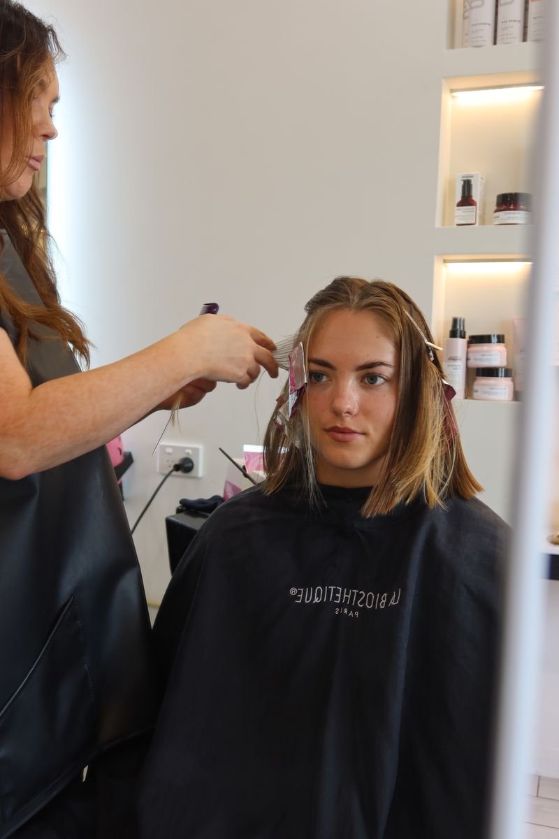 A woman getting her hair dyed or styled at a salon, sitting in a chair with a salon cape on, while a stylist works on her hair.