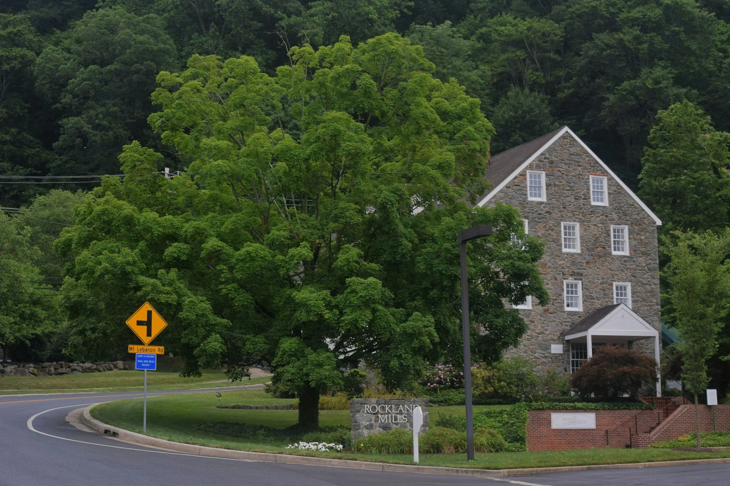 A street corner with a large green tree, a stone building labeled 'Rockland Mills', and a street sign for Mount Lebanon Road, surrounded by lush greenery and hillside trees.