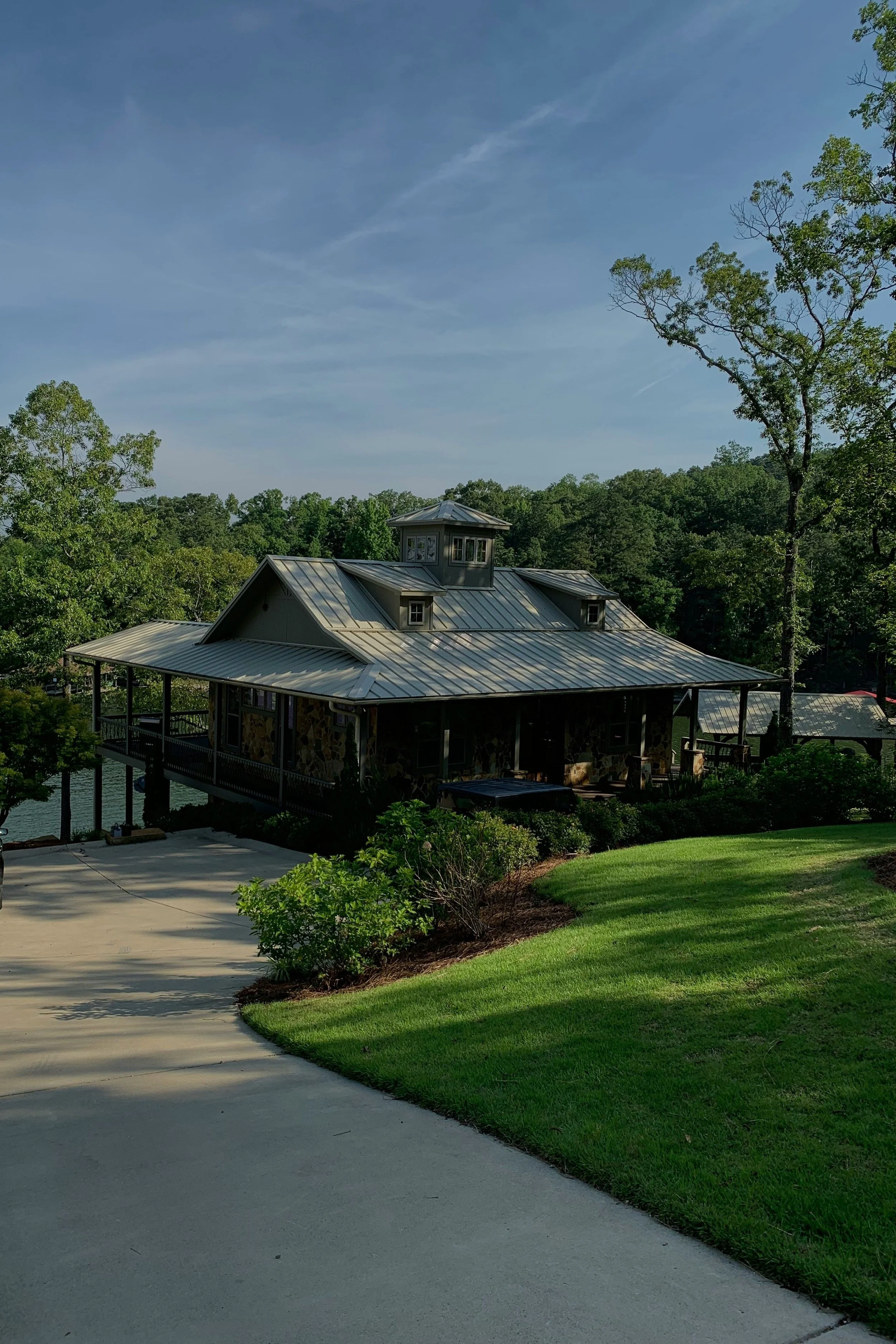 A house with a metal roof surrounded by trees, with a driveway and lawn in the foreground and a body of water partially visible behind the house.