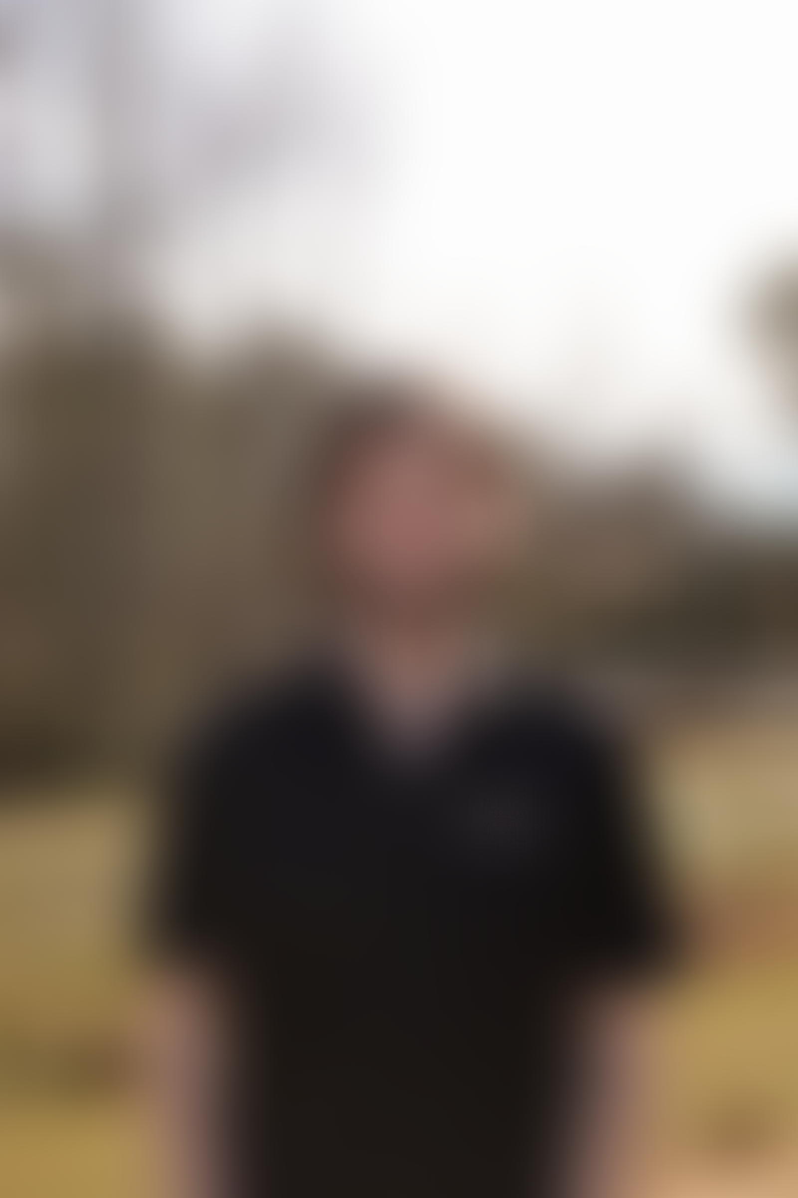 Blurry image of a man standing outdoors, possibly in a park or garden, with trees and a cloudy sky in the background.
