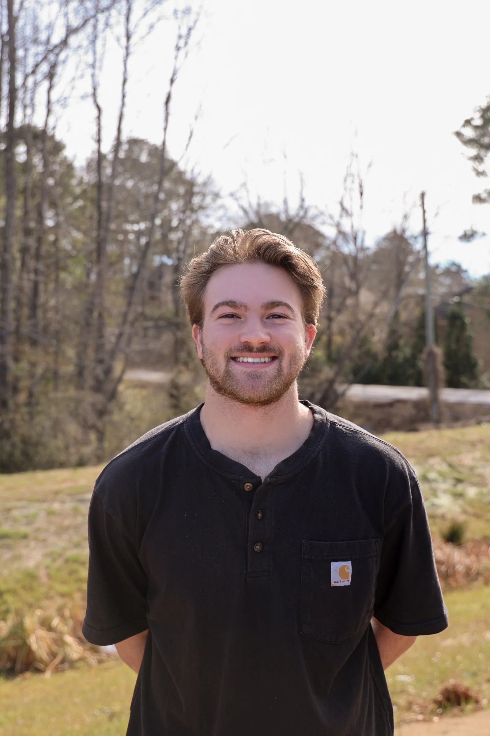 A young man with light brown hair and a beard smiling outdoors on a partly cloudy day, wearing a black Carhartt shirt, with trees and open grass in the background.