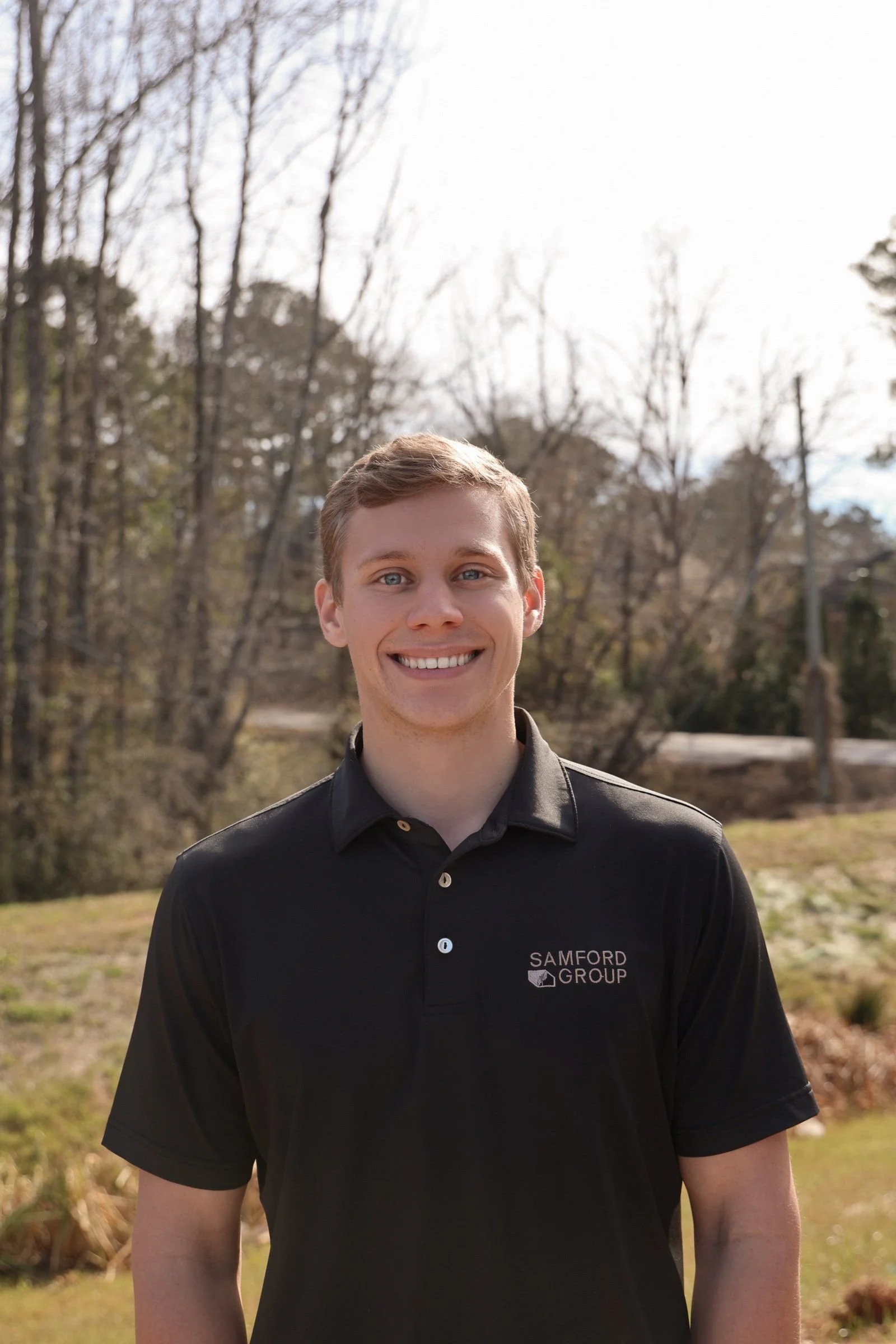 Josh Mura, President of Samford Group, smiling outdoors wearing a black polo shirt, with trees and a partly cloudy sky in the background.