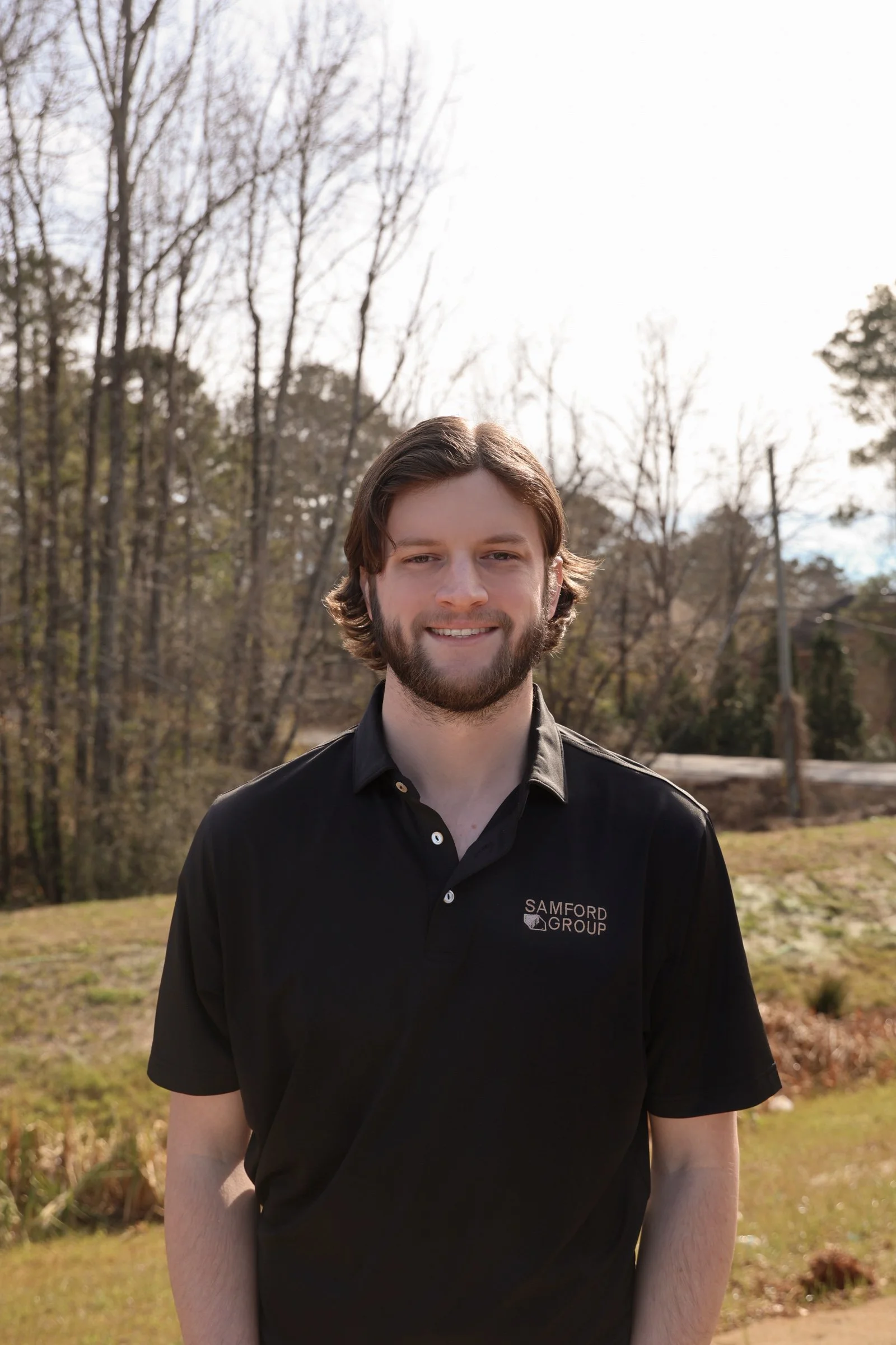 A young man with brown hair and a beard smiling outdoors, wearing a black polo shirt with 'Samford Group' logo, in a natural setting with trees and grass.