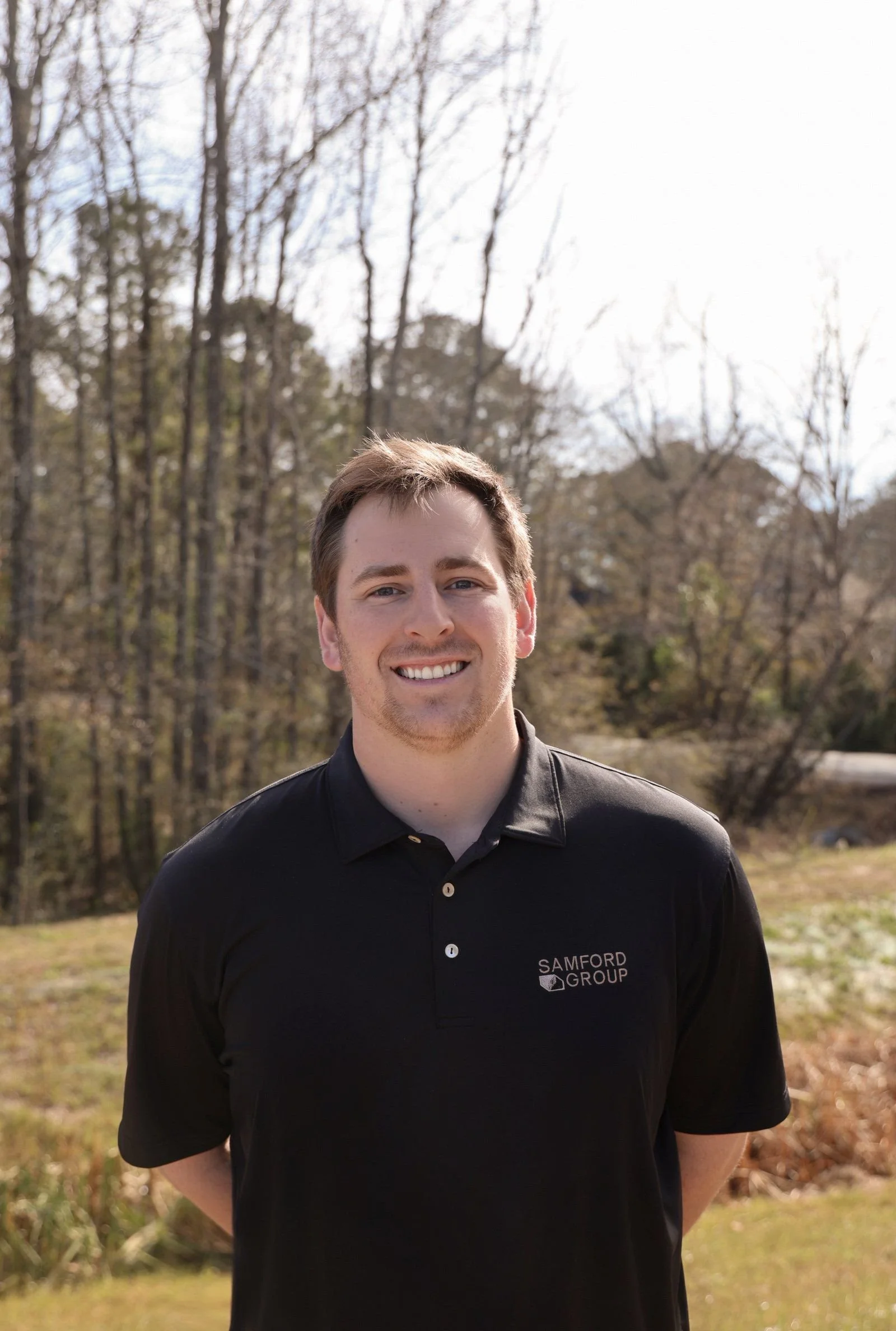 Hayes Eiford, founder of Samford Group, smiling outdoors in front of a background of leafless trees and a clear sky, wearing a black polo shirt.