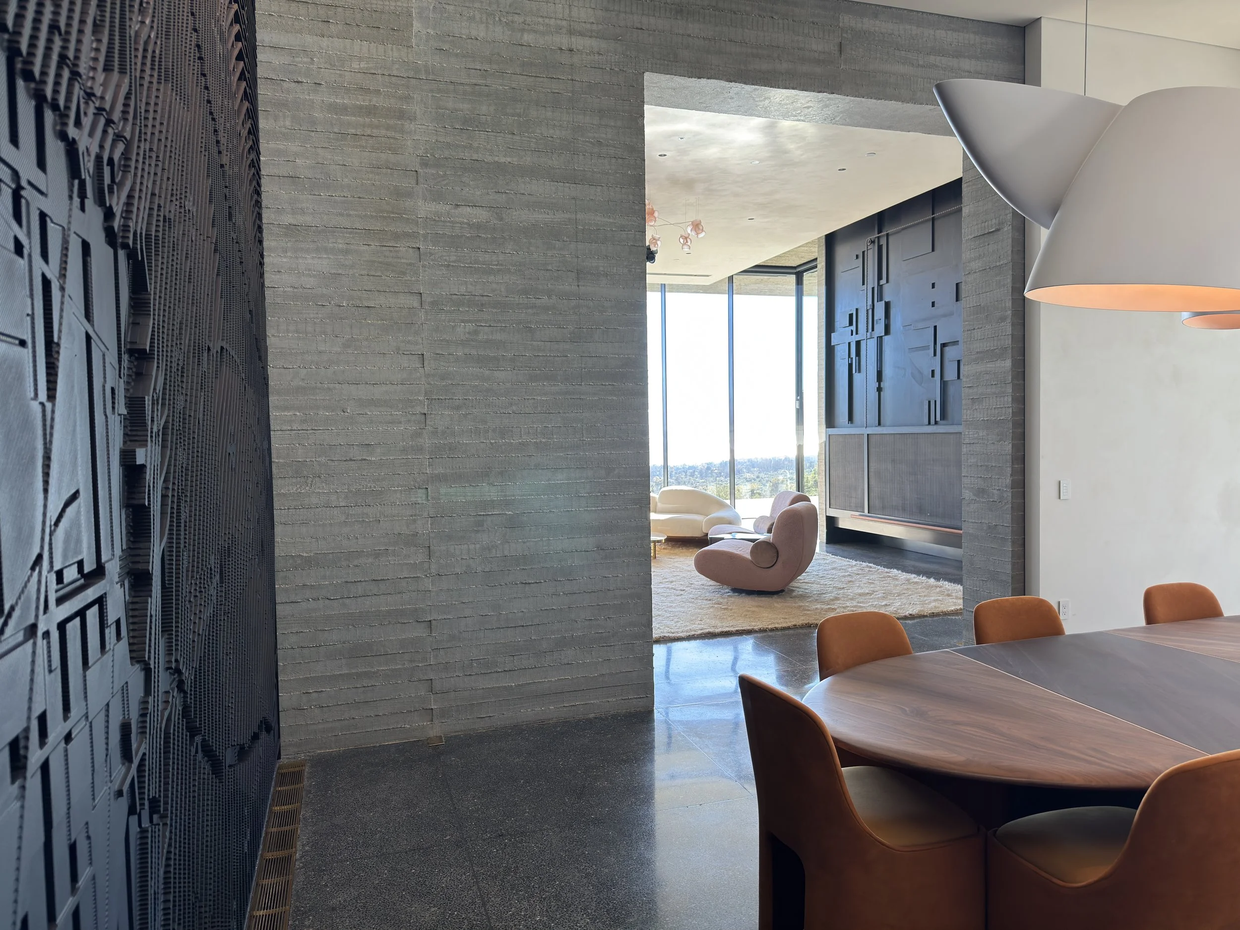 Interior view of a modern living and dining area with a round wooden table, orange upholstered chairs, a textured gray wall, abstract white and pink chairs, large windows, and a view of the cityscape.