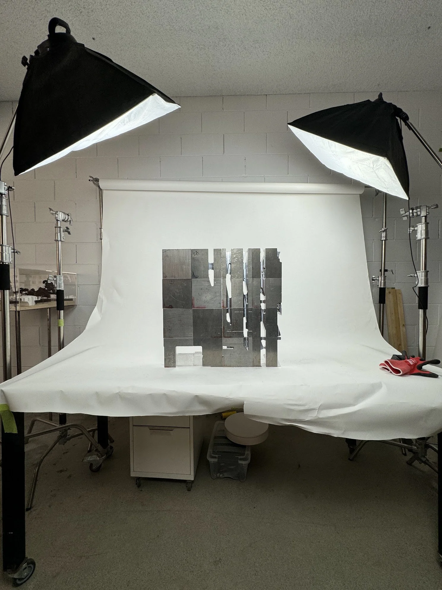 Photography studio setup with two softbox lights directed at a white backdrop. The backdrop is draped over a table, with metallic sculptural blocks arranged on it.