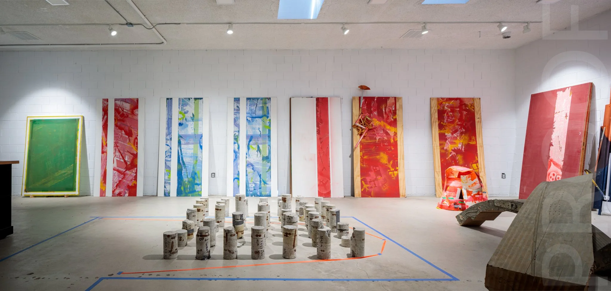 Art gallery with colorful abstract paintings on white brick wall, sculpture made of concrete cylinders, and art objects on the floor.