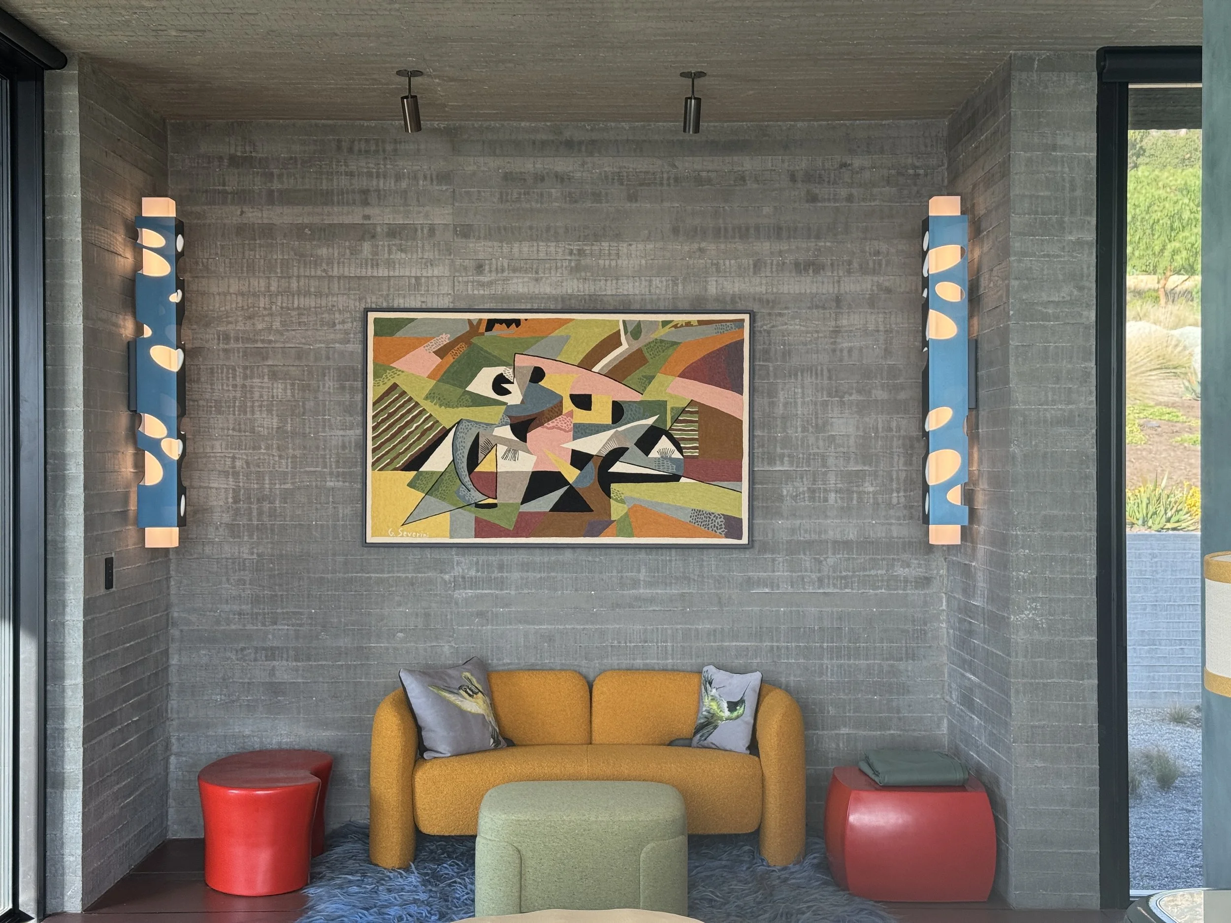 Modern living room with gray brick walls, colorful abstract artwork, yellow sofa with decorative pillows, two tall blue wall sconces, and red side tables.