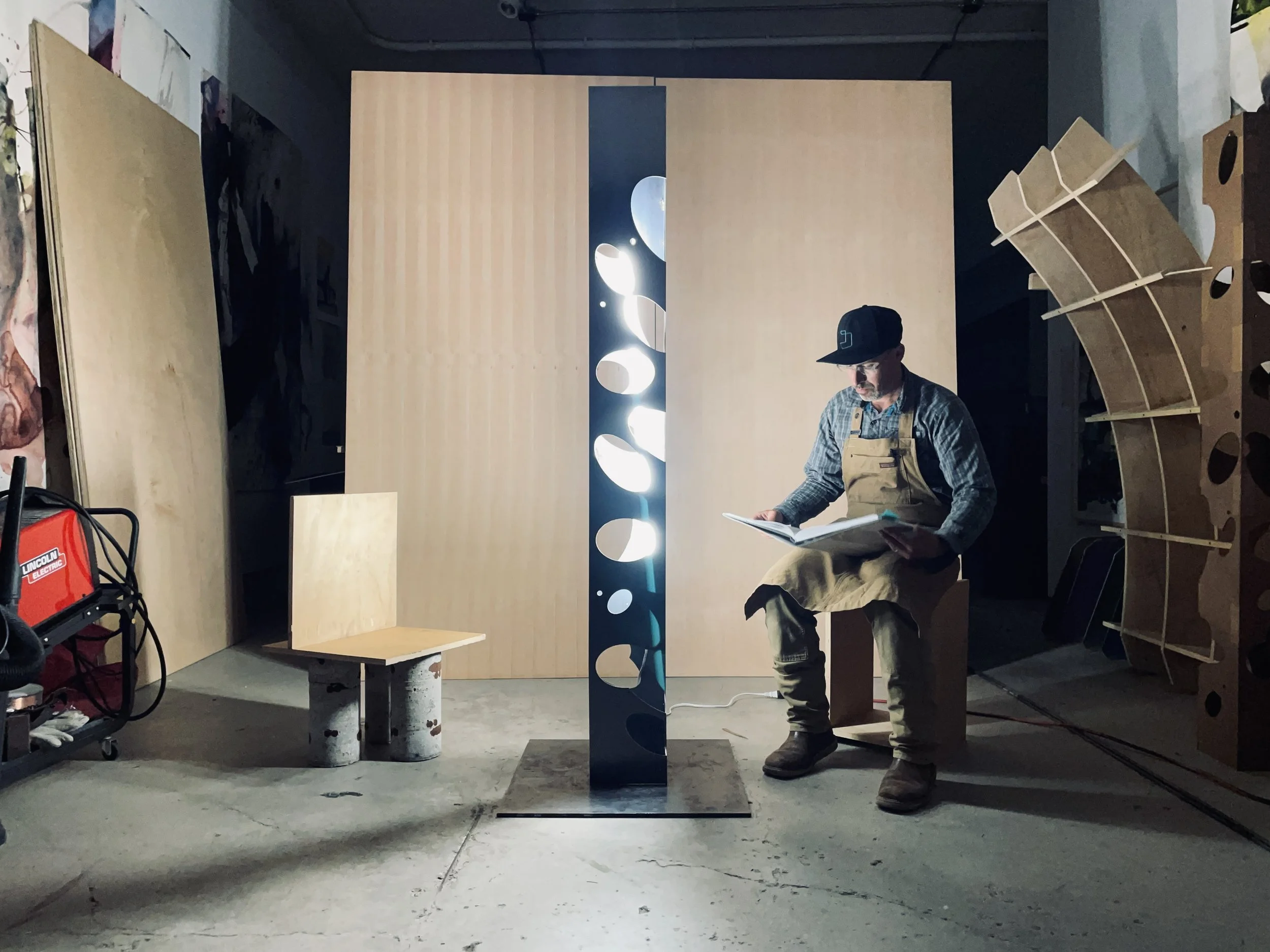 Man sitting on a wooden block, reading a book, surrounded by various wooden and metal structures in a workshop with a large wooden panel and an illuminated decorative piece in the background.
