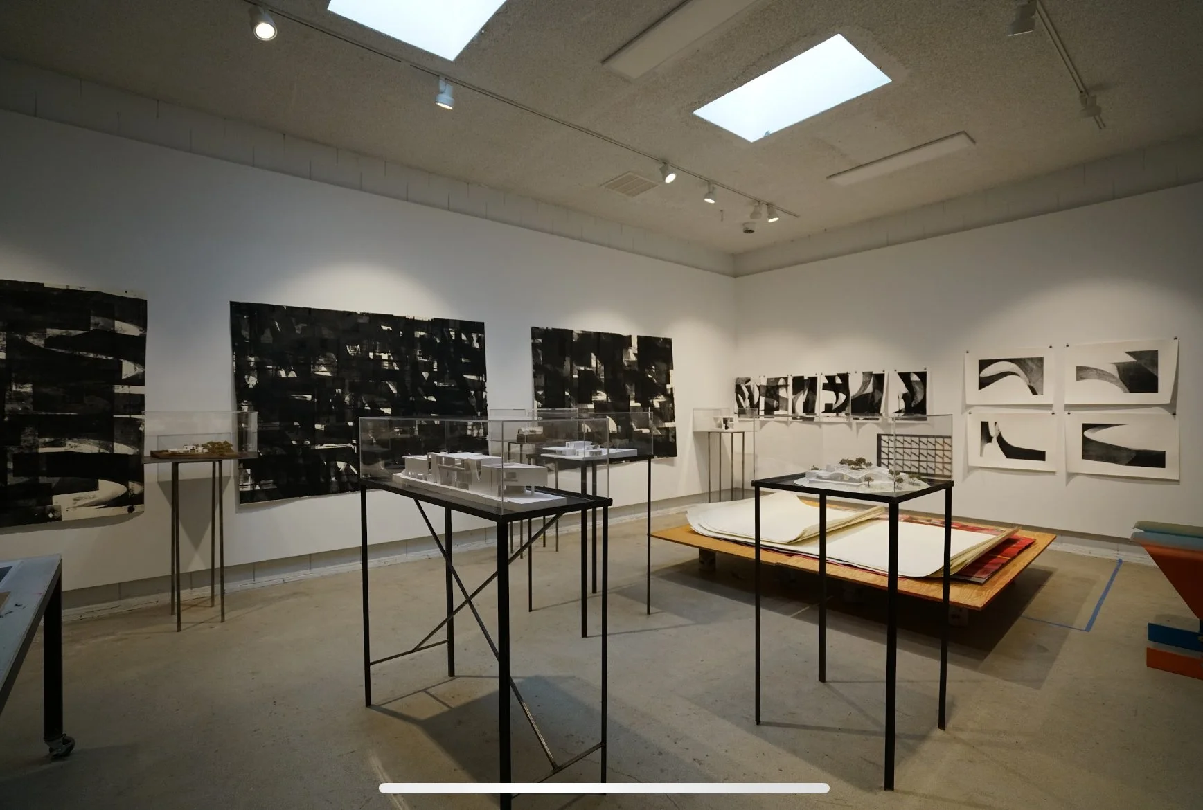 An art gallery with black and white abstract artwork on the walls, display tables with models and artifacts, and a large open sketchbook on a platform in the center.